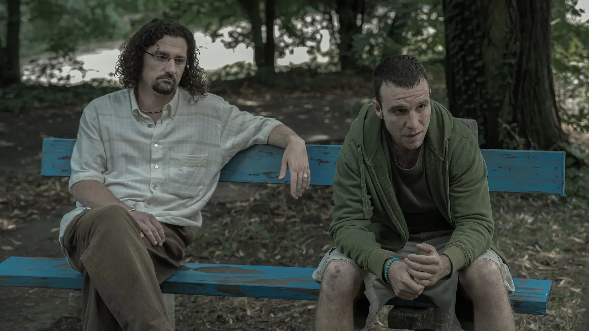 Two men sitting on a blue bench in a natural setting one is looking condescendingly at the other, while the second is staring desperately.