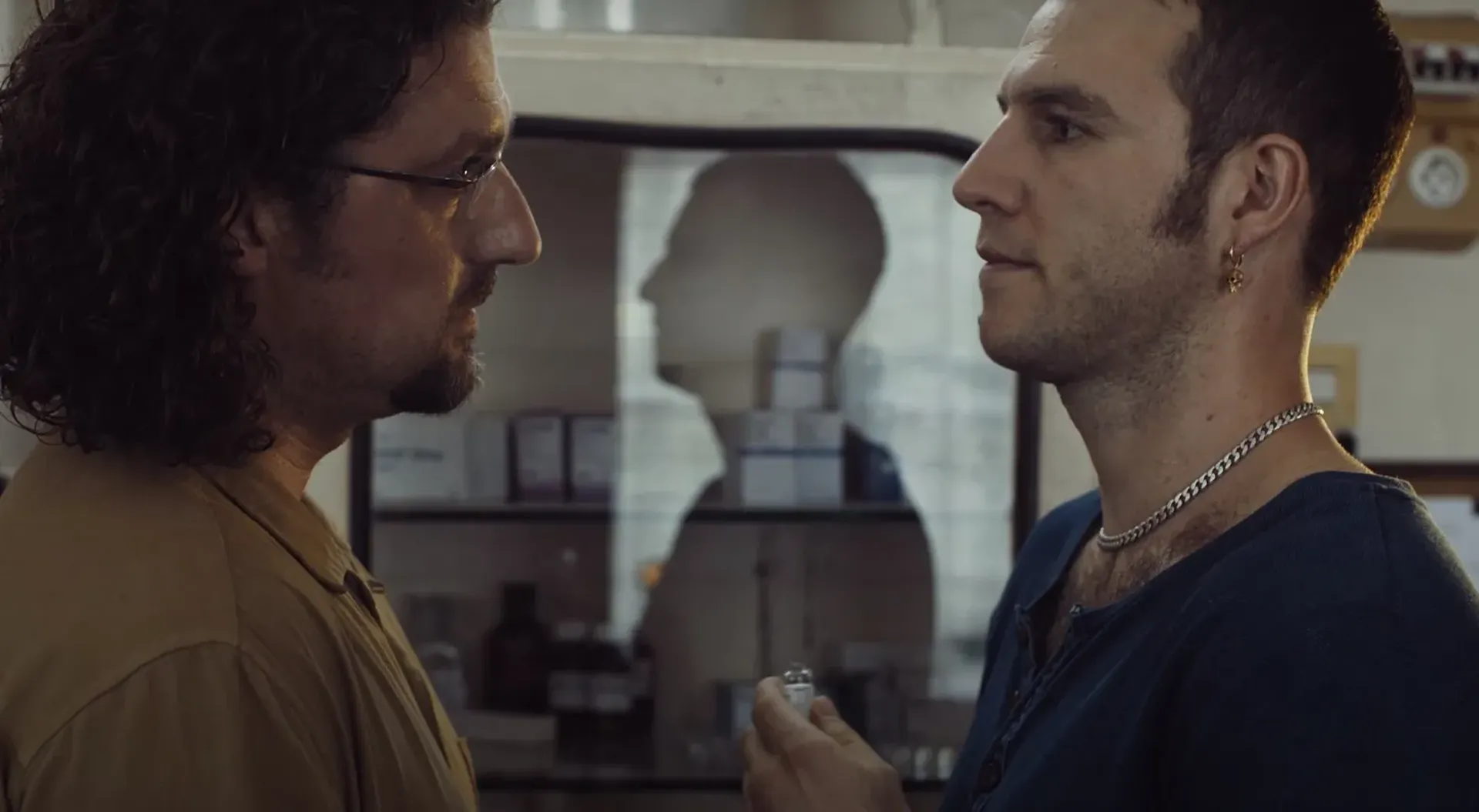 Two men staring at each other one with long curly hair, the other with short hair and stuble holding a vile.