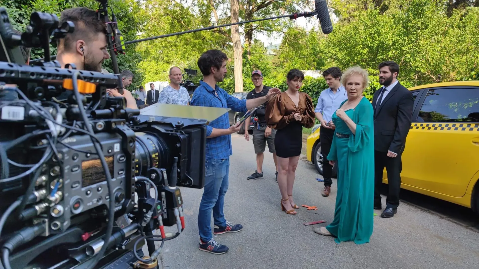 Film crew recording an outdoor street scene with actors standing beside a yellow car, professional camera rig in the foreground, captured with flash lighting.