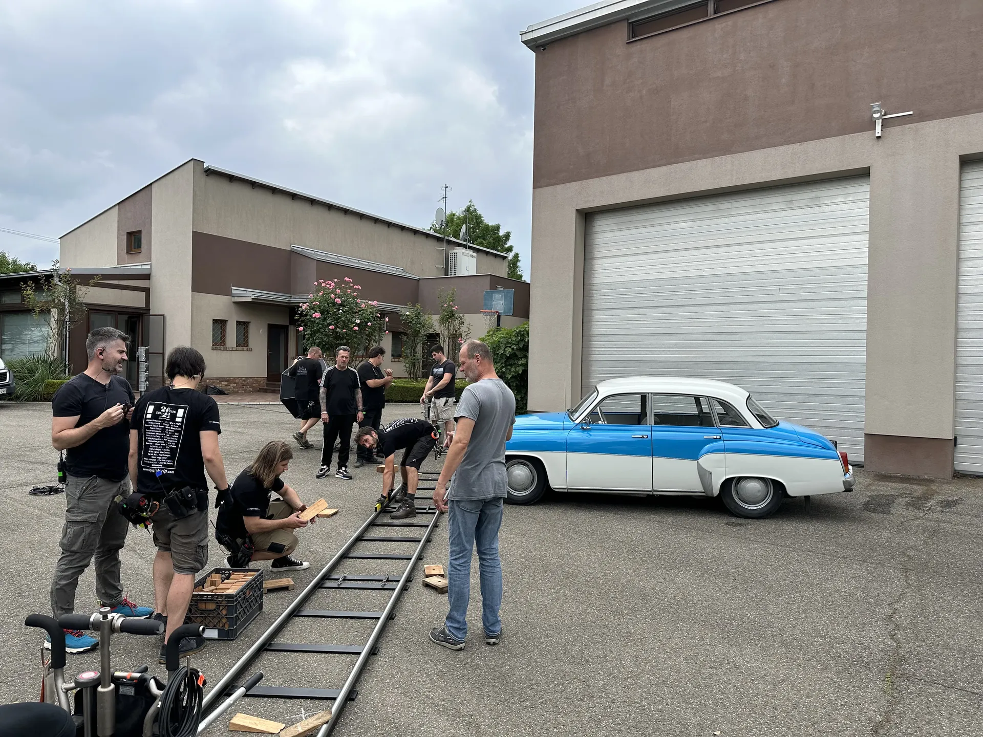 Film crew setting up camera dolly tracks in an industrial courtyard with a vintage blue-and-white car parked nearby, photographed with flash lighting.