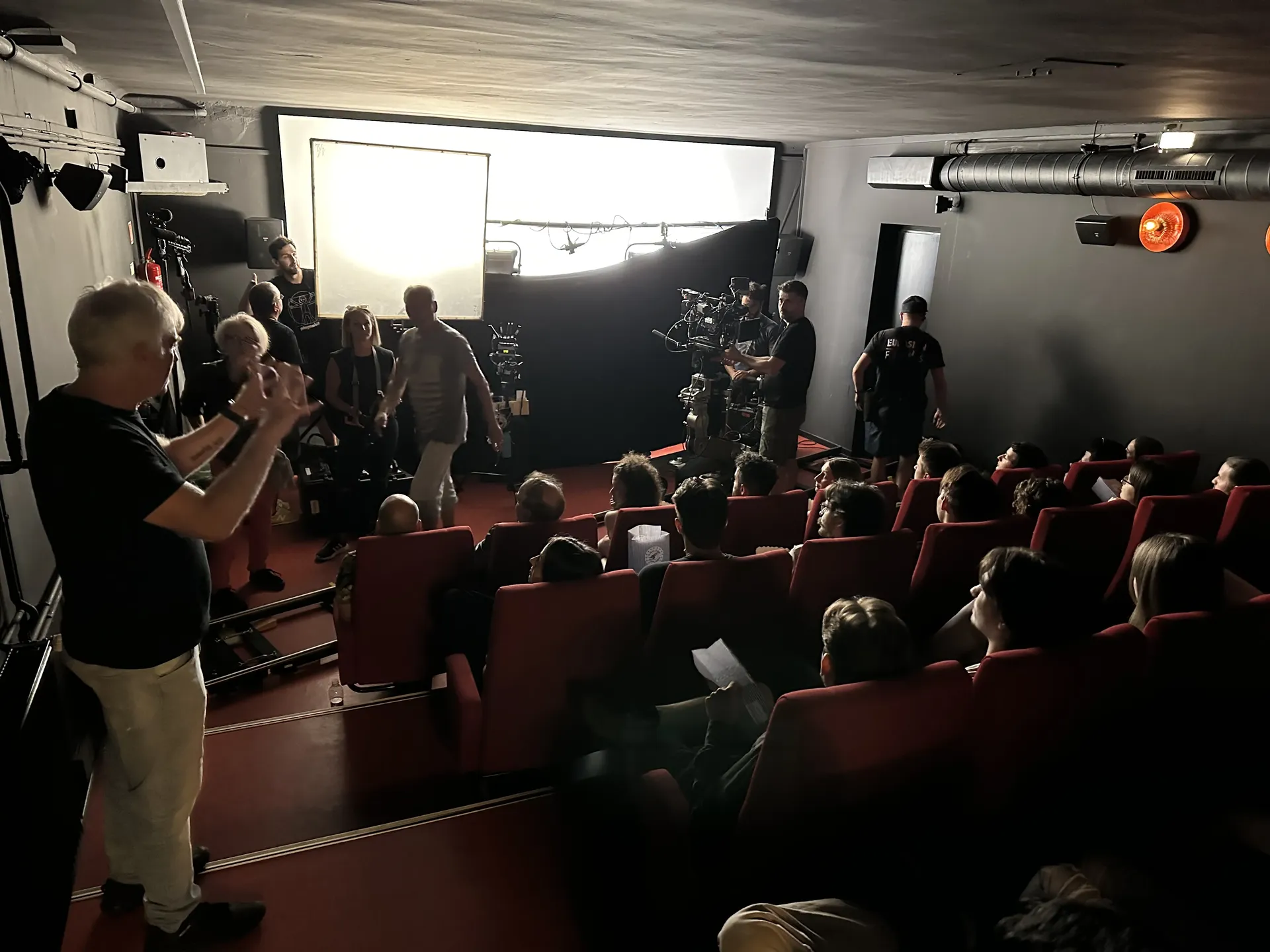 Film crew shooting a scene inside a small cinema with actors seated in red chairs and lighting equipment set up near the screen, photographed with flash lighting.