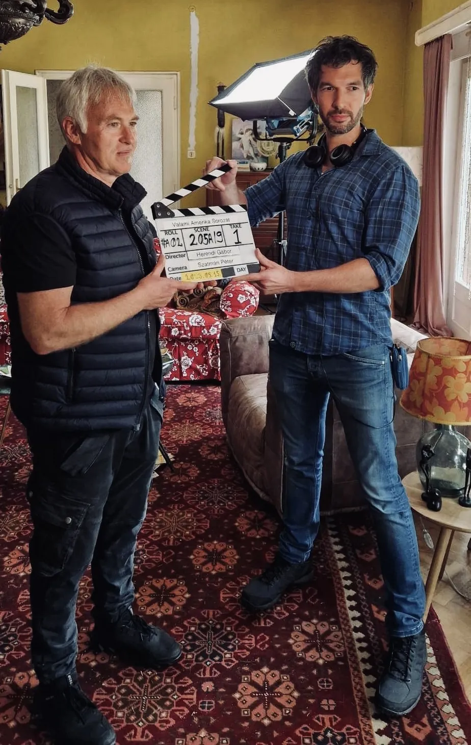 Two crew members standing in a furnished interior holding a film slate for a production, with patterned carpet and set decor visible, photographed with flash lighting.