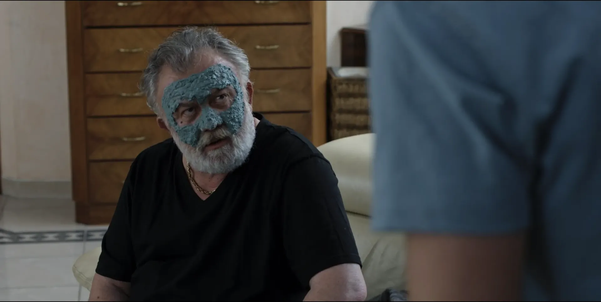 Older man sitting indoors with a textured blue facial mask covering the upper part of his face, looking toward someone off-screen, photographed with flash lighting.