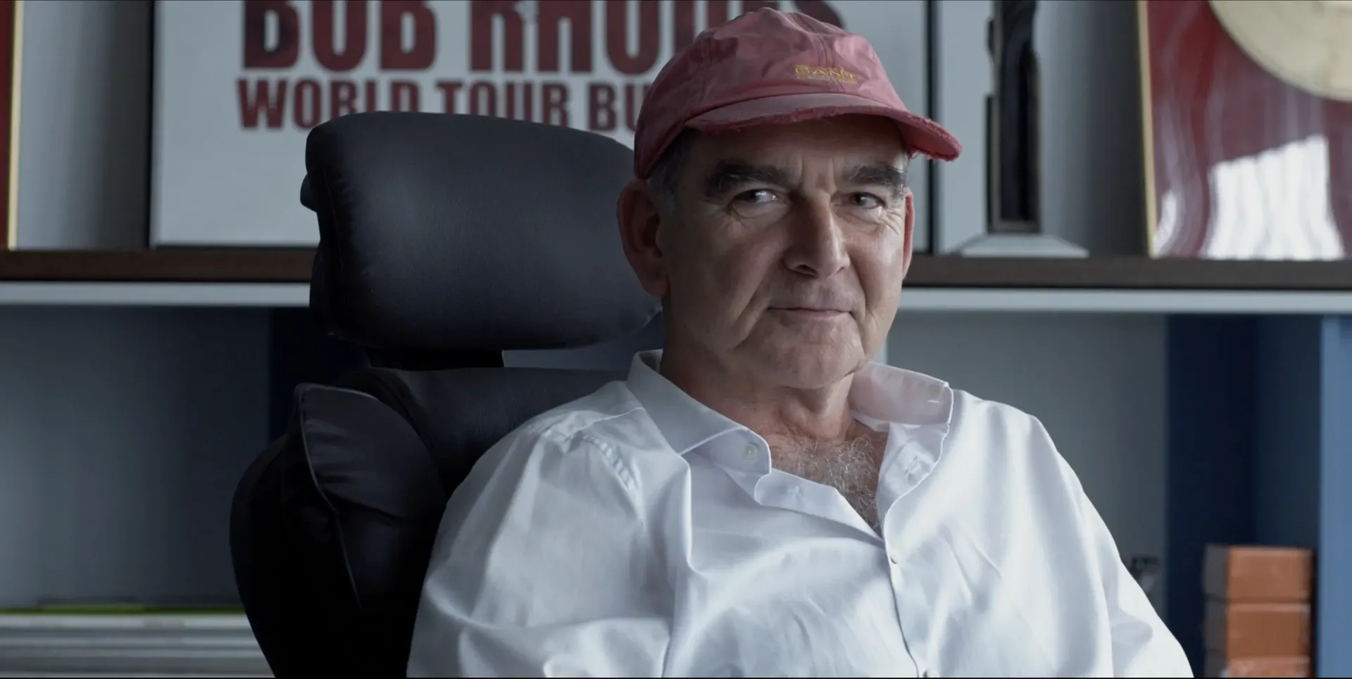 Man in a pink cap and white shirt sitting in a black office chair, looking toward the camera with shelves and framed posters behind him, photographed with flash lighting.