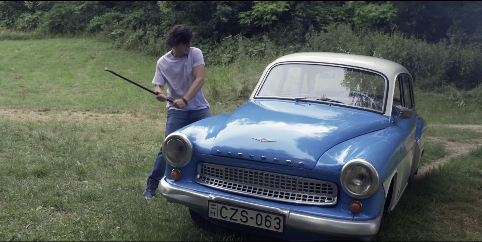 Man in a grassy field swinging a metal rod toward a vintage blue Wartburg car, with trees in the background and the scene captured with flash lighting.