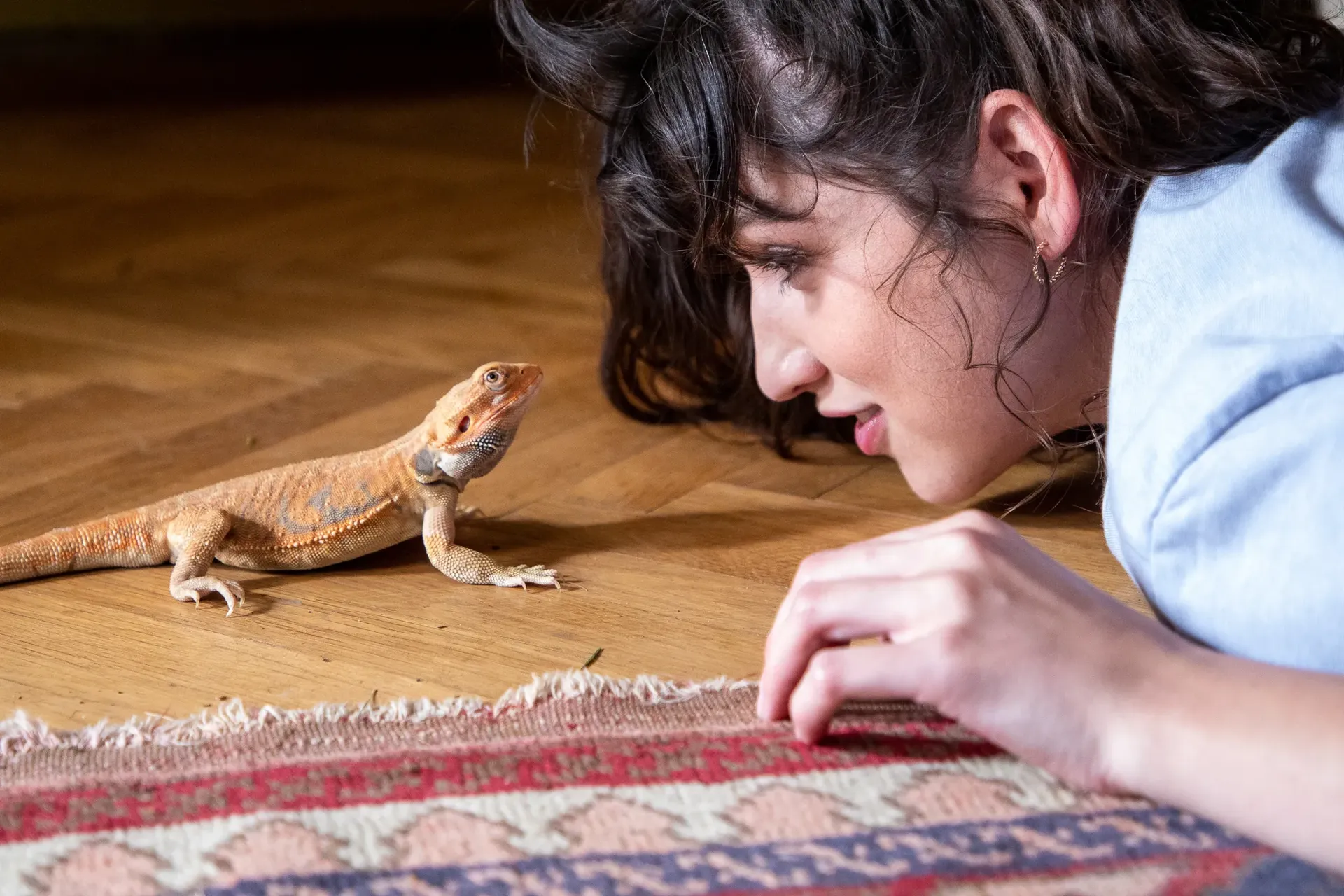 Woman lying on the floor face-to-face with a small orange lizard near a patterned rug, captured with bright flash lighting for sharp detail.
