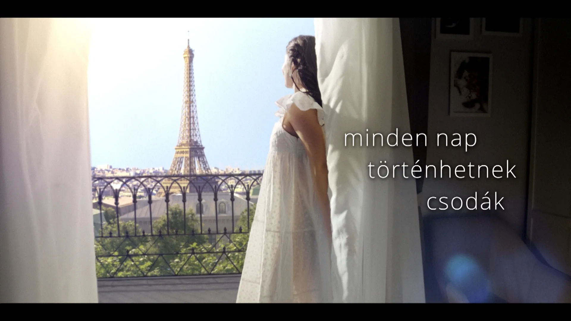 A woman in a white nightgown stands by an open balcony door, looking toward the Eiffel Tower in bright daylight, with soft curtains framing the scene and Hungarian text reading “minden nap történhetnének csodák” on the right side.