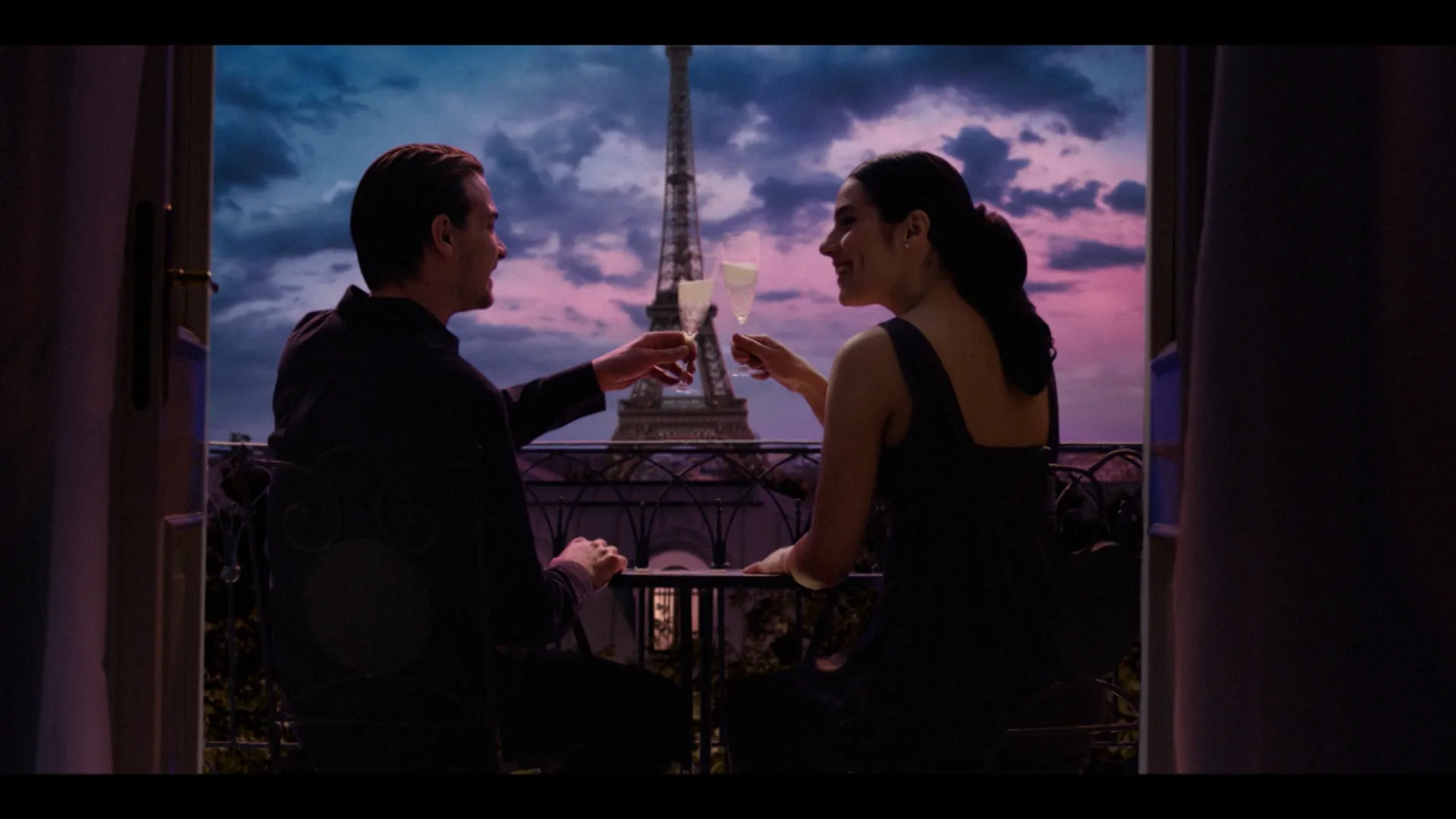 A man and a woman sit on a balcony at dusk, clinking champagne glasses with the Eiffel Tower glowing in the background.