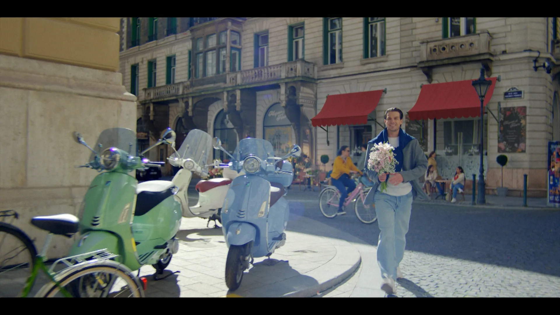 A smiling man walks down a sunlit European street holding a bouquet of flowers, passing parked pastel-colored scooters as cyclists and café patrons move in the background.