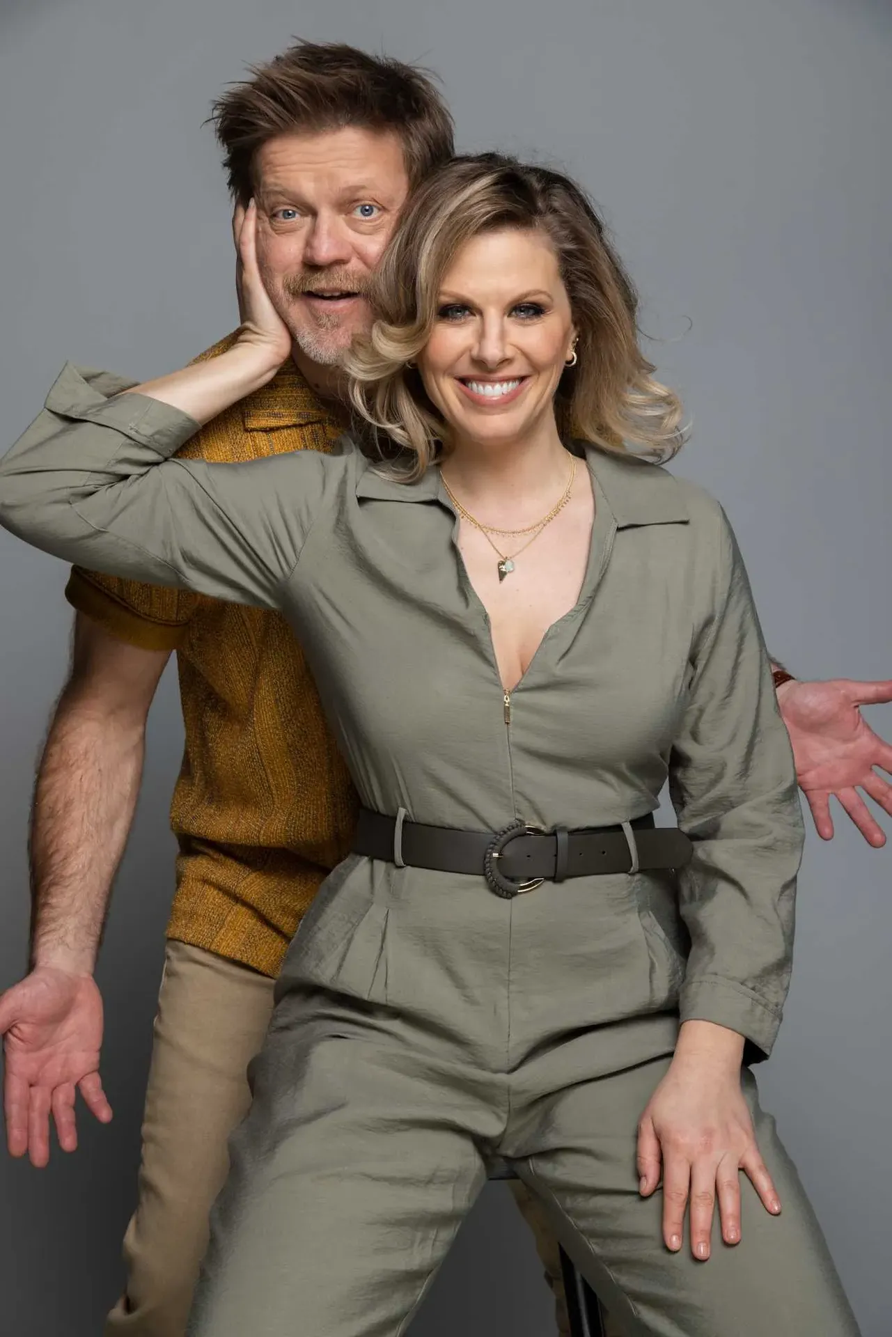 A smiling couple poses playfully together in a studio, with the woman in front and the man leaning from behind her.