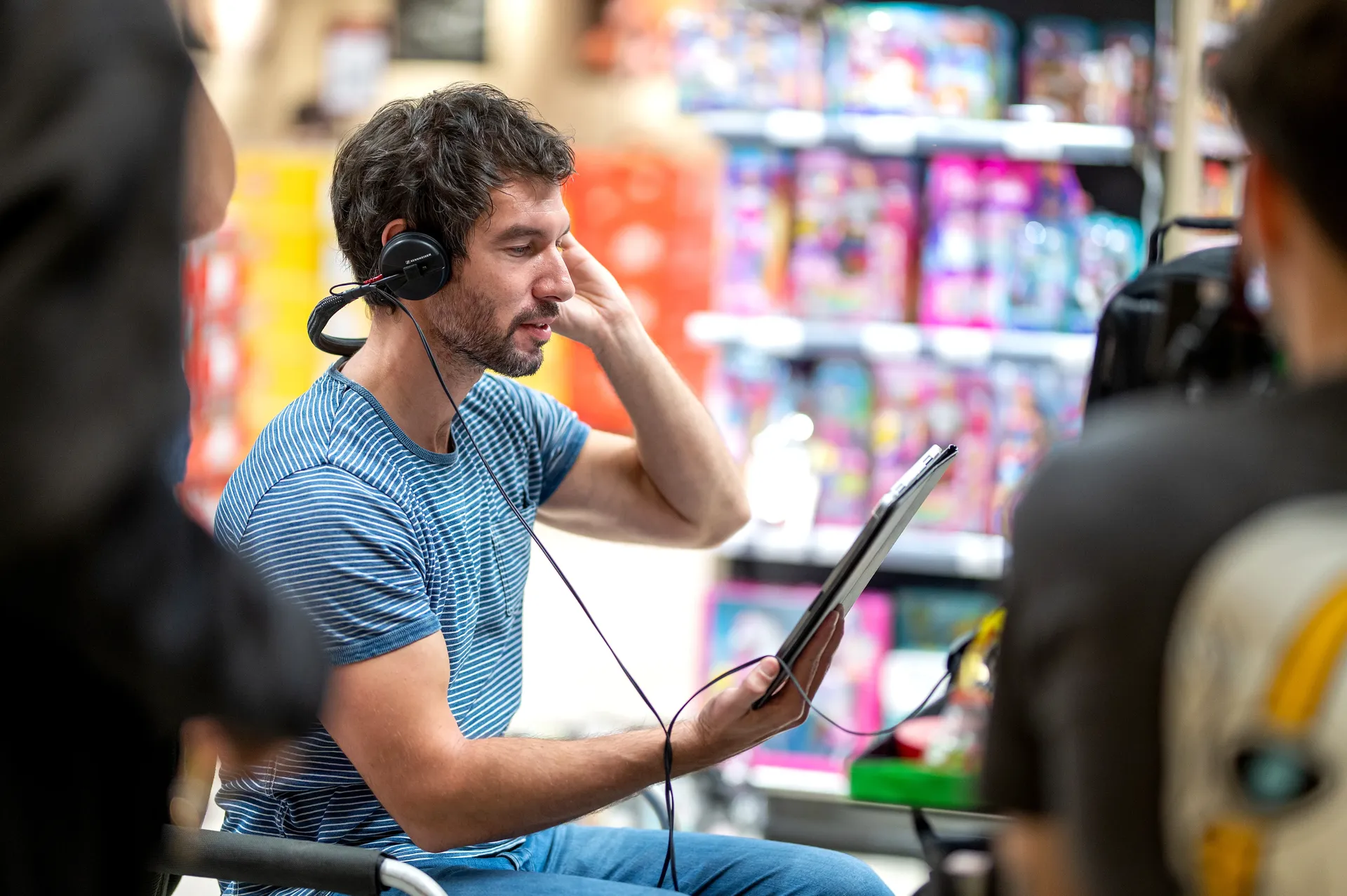 A crew member with headphones reviews a tablet on set in a brightly lit store aisle.