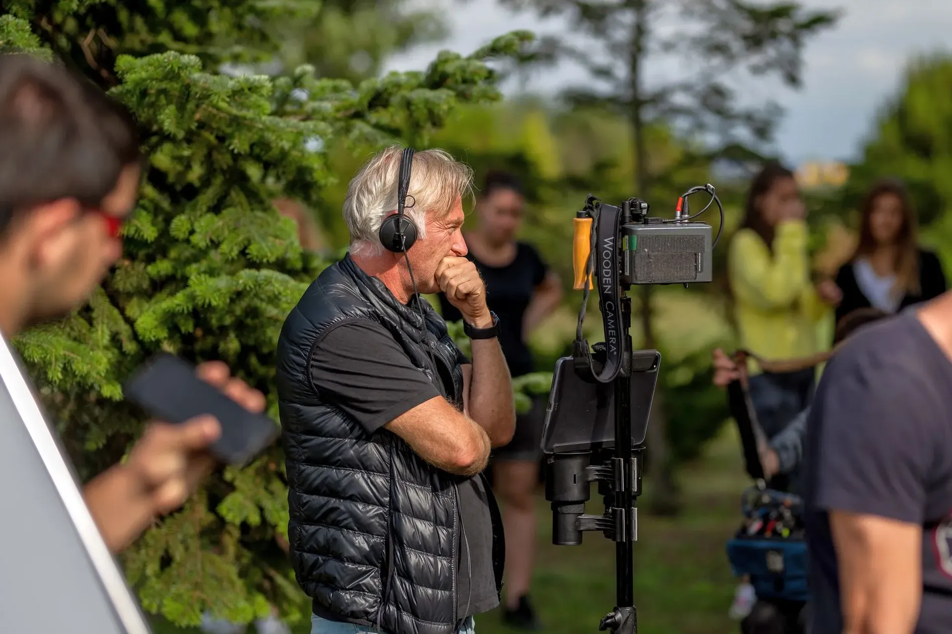 A director watches a monitor outdoors during filming, surrounded by crew members and equipment.