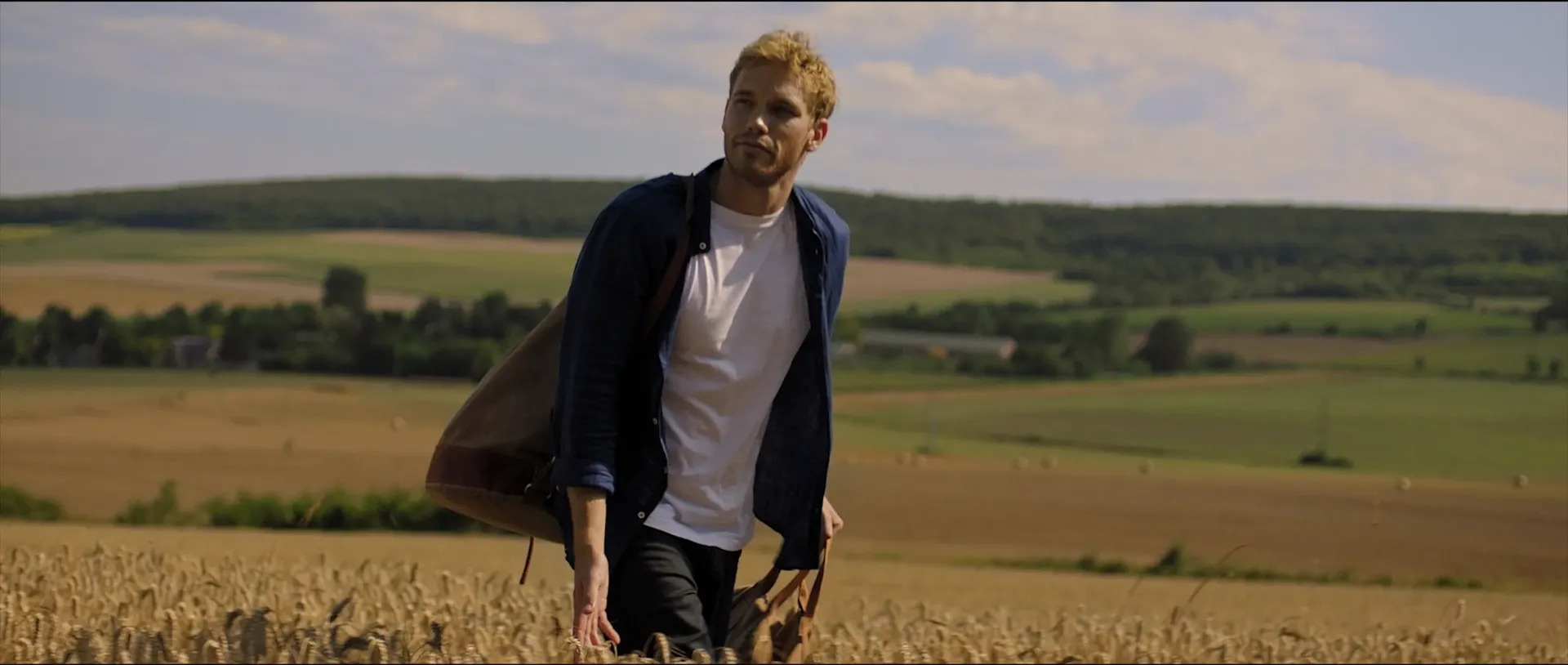 A man walks through a golden wheat field with a bag over his shoulder, surrounded by rolling green hills under a bright sky.
