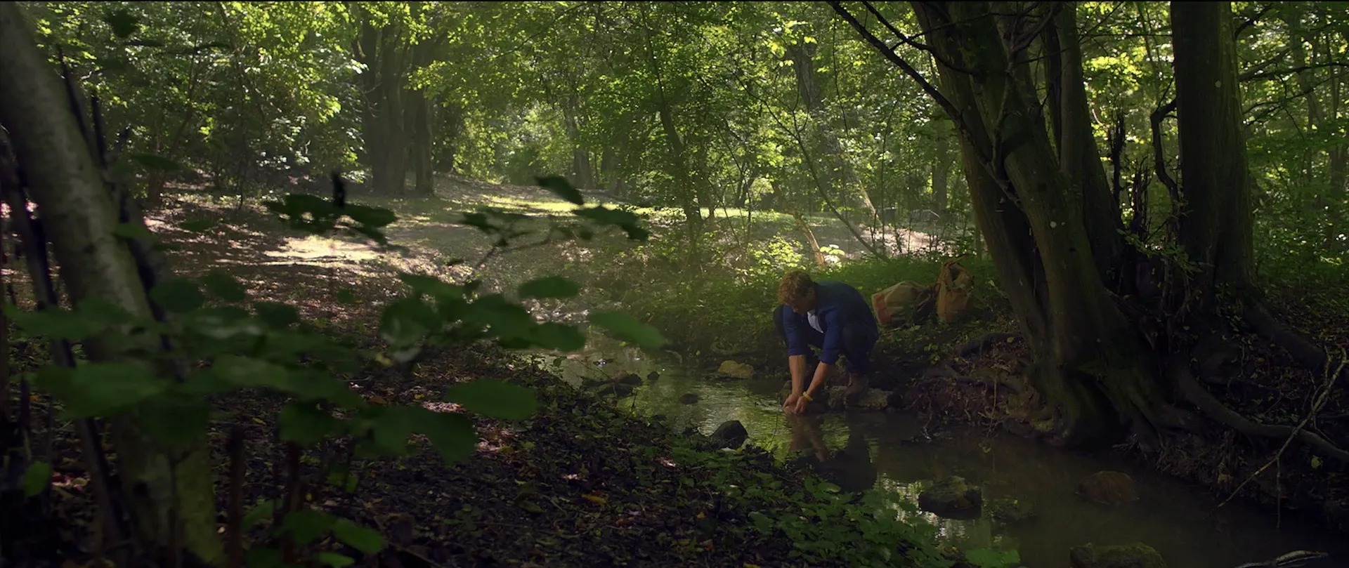 A man kneels by a small forest stream, cupping water in his hands amid lush green trees and filtered sunlight.