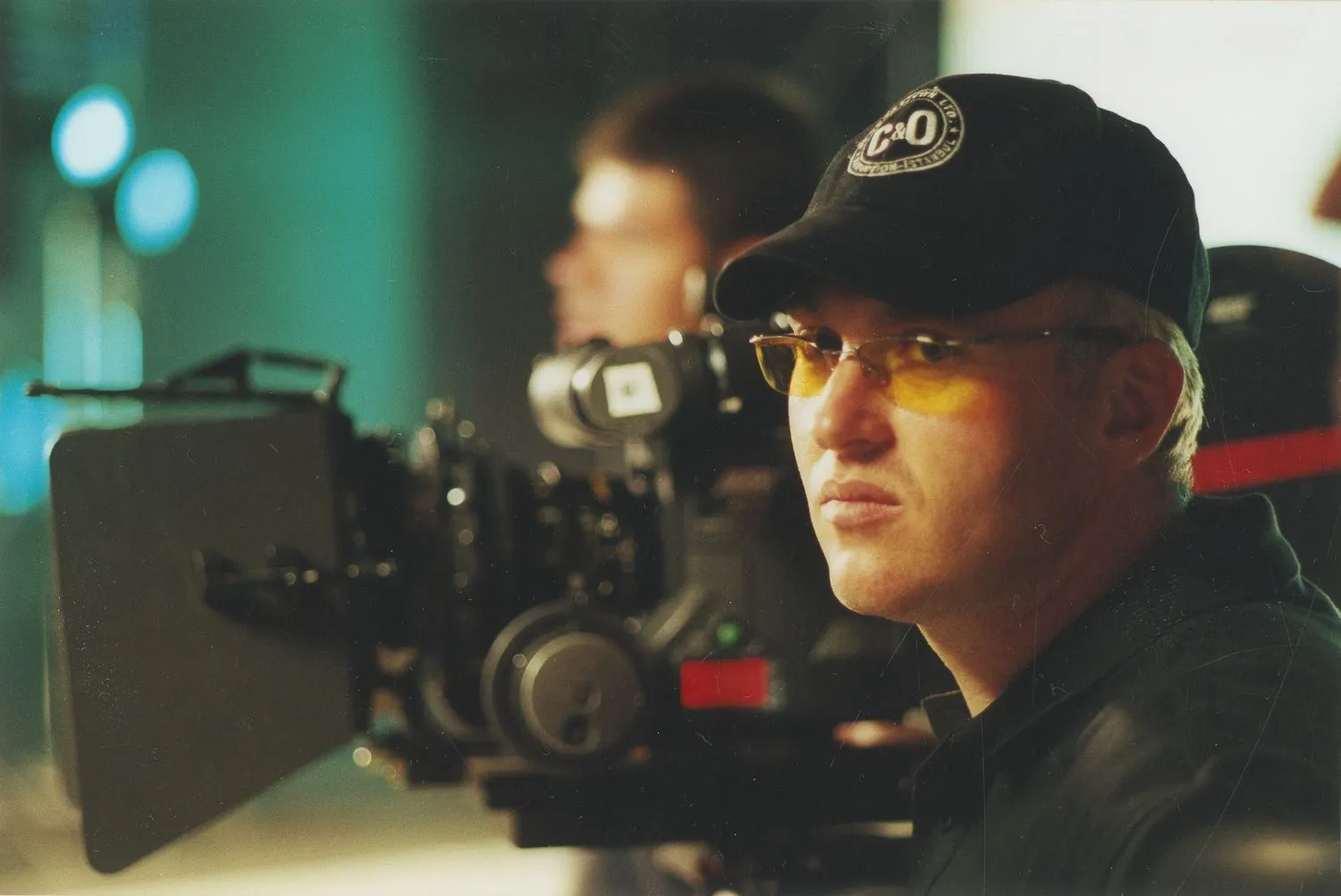 A focused camera operator wearing a cap and tinted glasses looks through a professional film camera during a shoot.