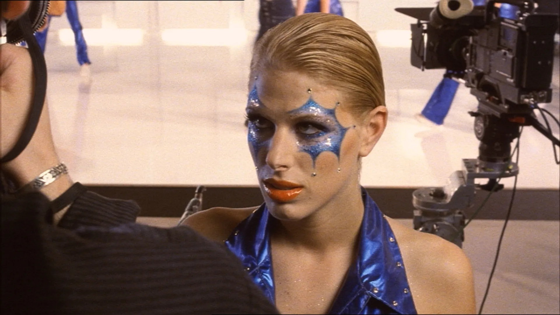 A woman in bold blue stage makeup and a shiny blue outfit looks intensely toward someone off-camera on a studio set.