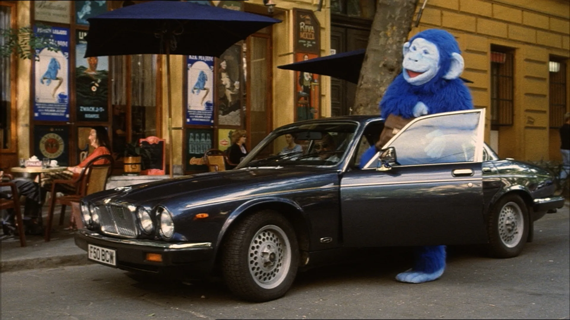 A person in a bright blue monkey costume steps out of a parked black classic car on a city street near a café.