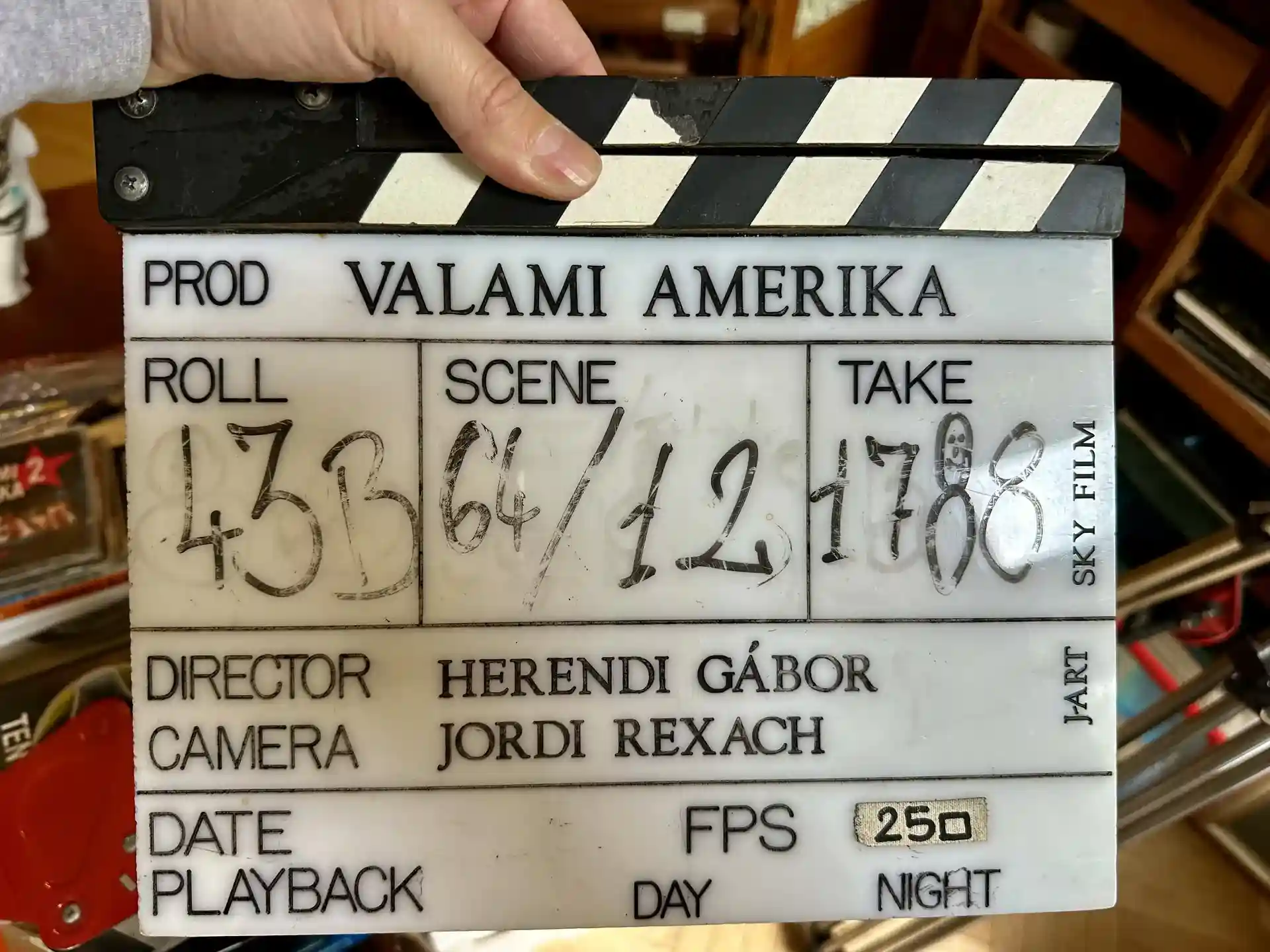 A film slate labeled Valami Amerika is held up, showing roll, scene, and take information along with the director and camera credits.
