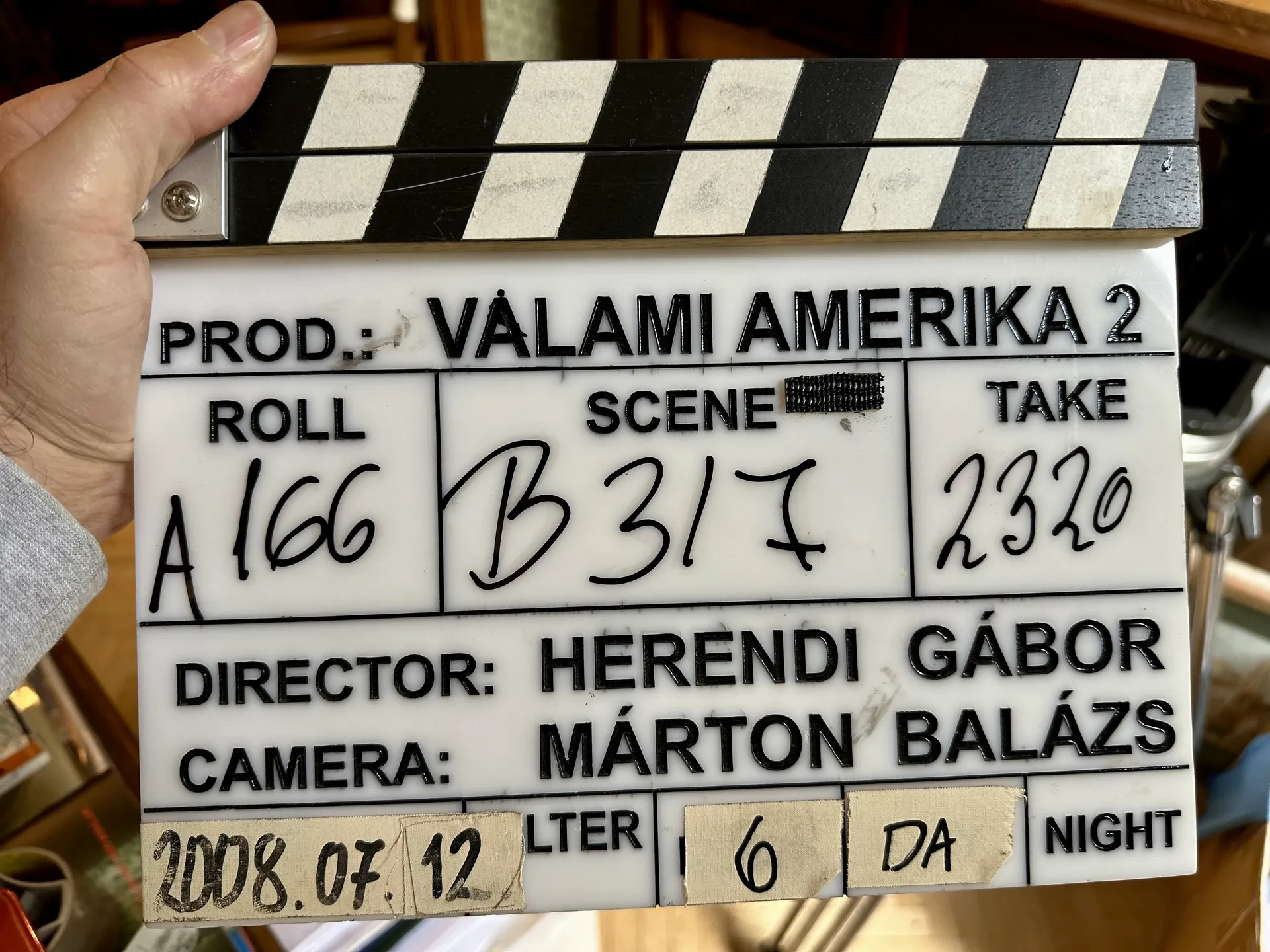 A film slate labeled Valami Amerika 2 being held up indoors, showing roll A166, scene B317, take 2320, with the director and camera operator’s names written on it.