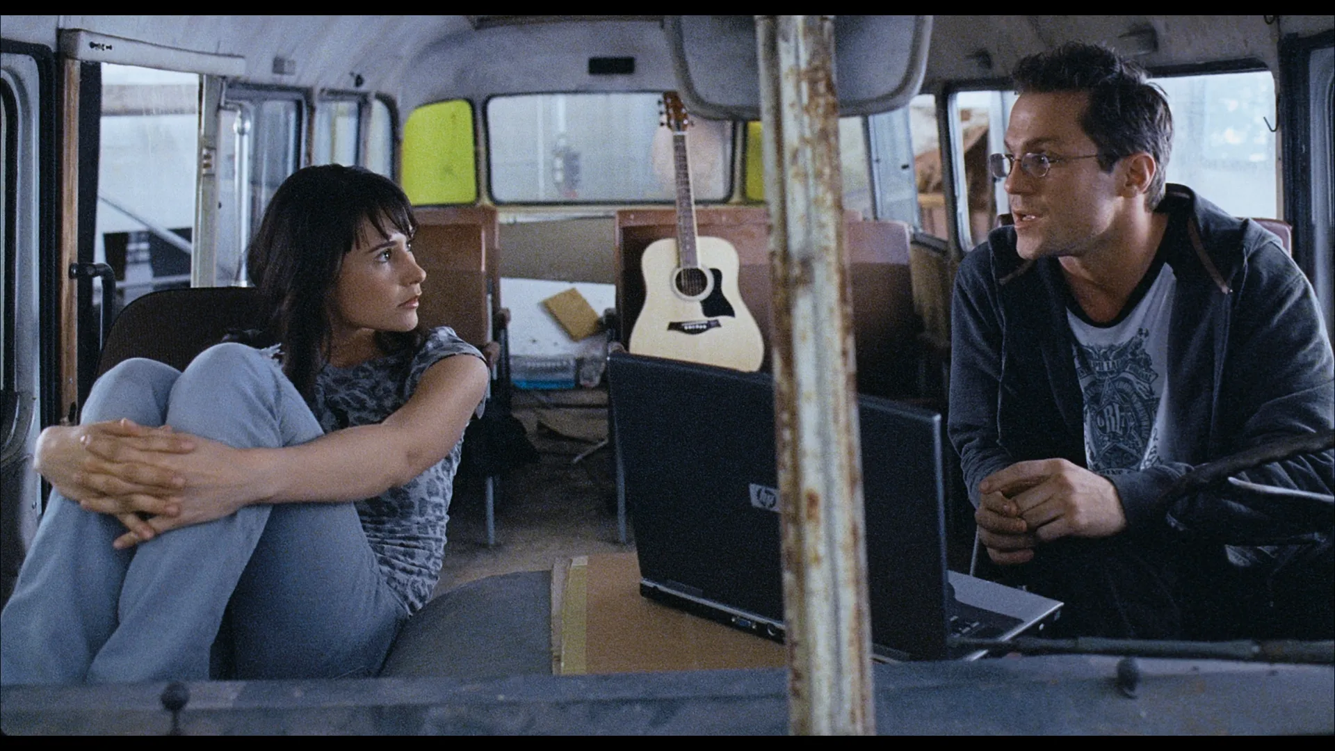 A woman sits curled up on a bus seat facing a man with glasses who is talking to her; an open laptop and an acoustic guitar are visible in the worn interior of the bus.
