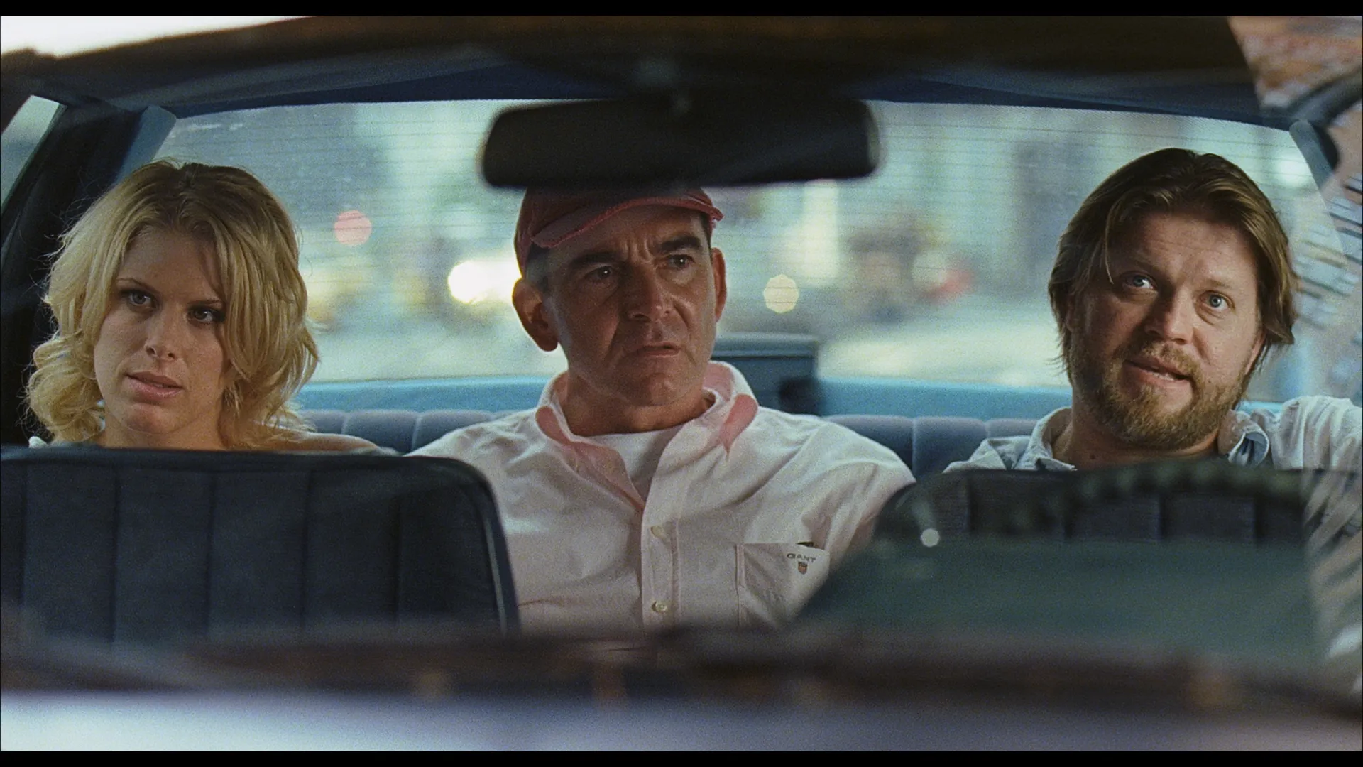Three people sit in the back seat of a car, a blonde woman on the left looking serious, a man in a pink shirt and cap in the middle appearing tense, and a bearded man on the right speaking with a slight smile.