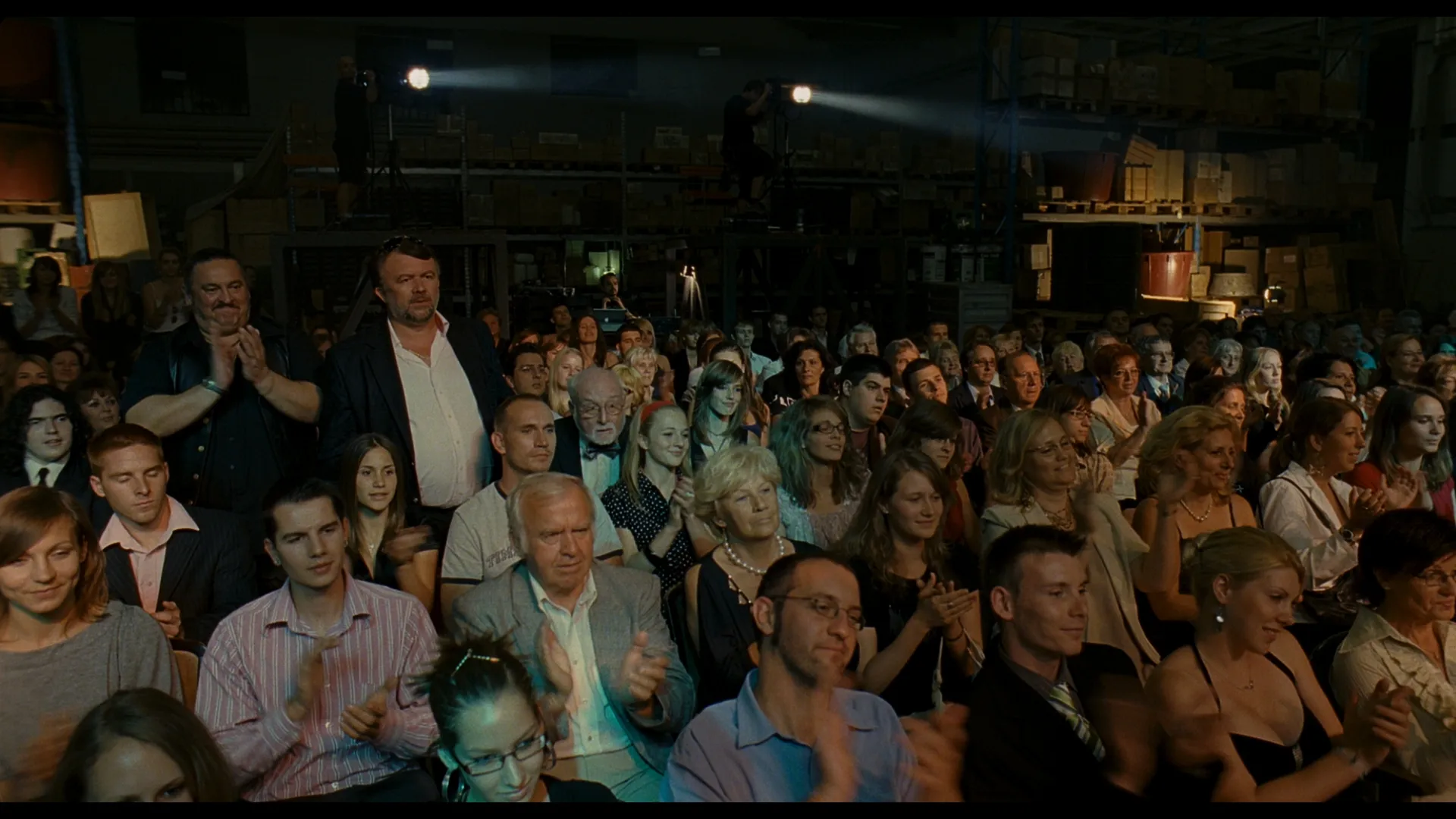 A large audience seated in a dimly lit venue applauding and smiling toward the stage, with some people standing while others cheer enthusiastically.