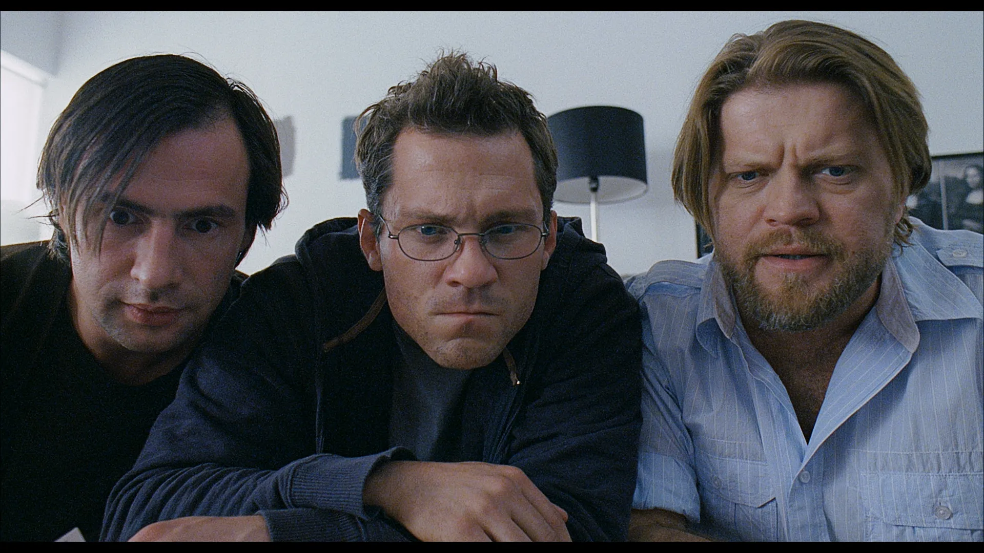 Three men lean close together and stare intensely forward, all wearing serious expressions in a brightly lit indoor setting.