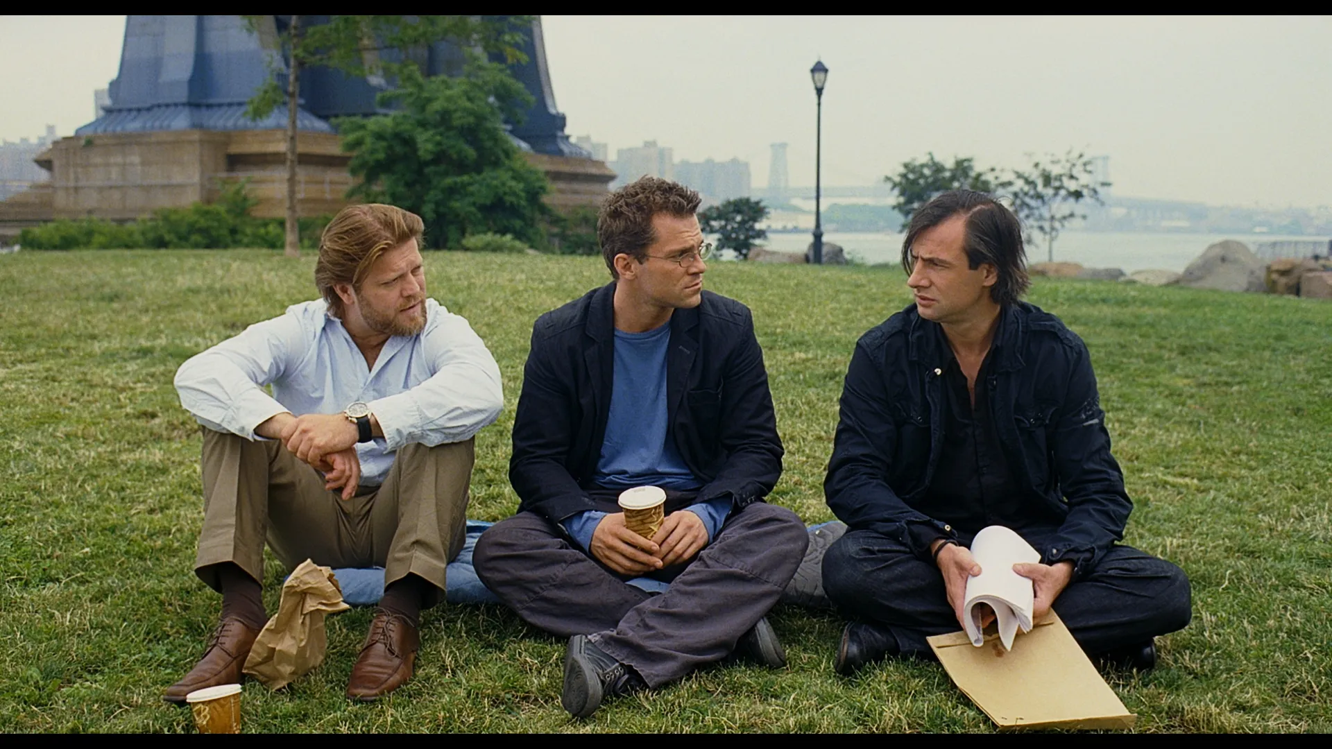 Three men sit on the grass outdoors, talking while holding drinks and food bags, with a foggy riverside and statue visible in the background.