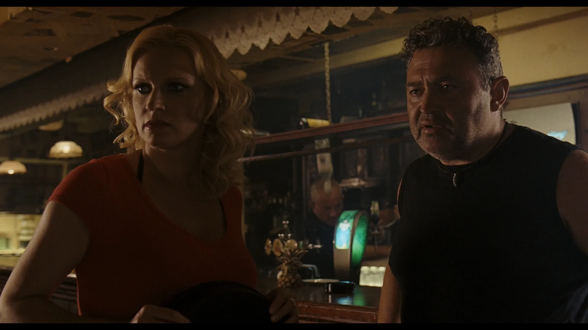 A woman in a red top and a man in a dark vest stand together in a dimly lit bar, looking concerned or startled.