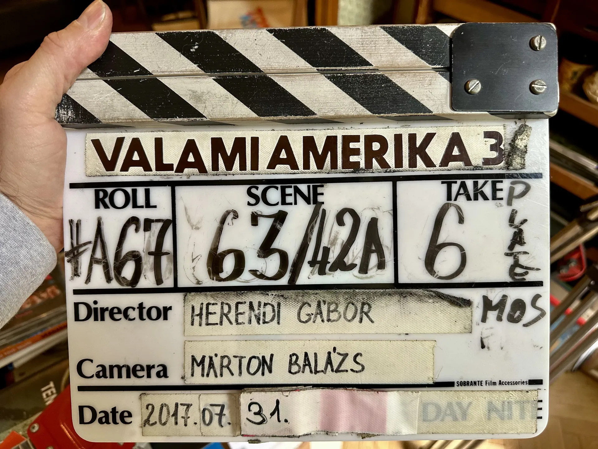 A film slate labeled Valami Amerika 3 held up by a hand, showing roll, scene, and take numbers along with the director and camera operator’s names.