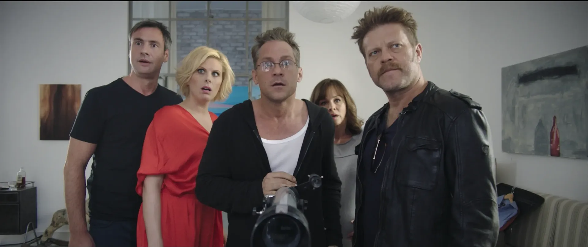 Five people stand close together indoors, staring ahead with surprised and tense expressions; the man in front is holding a large camera or device.