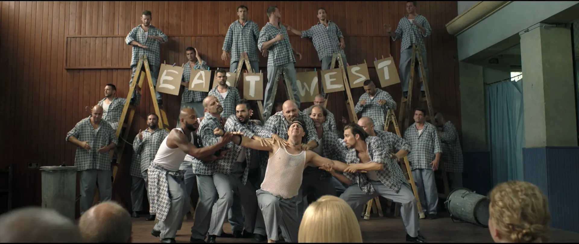 A large group of men dressed in matching checkered prison-style outfits perform a dramatic, choreographed scene in a hall, with some standing on ladders holding signs that read EAST and WEST while others grapple with a man posed theatrically at center stag