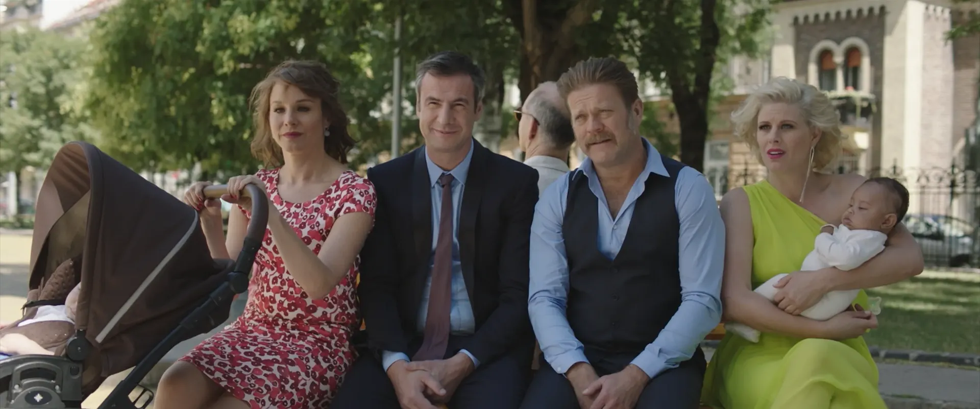 Four adults sit on a park bench in a sunny setting, with one woman pushing a stroller on the left and another woman on the right holding a sleeping baby, while the two men in the middle smile calmly.
