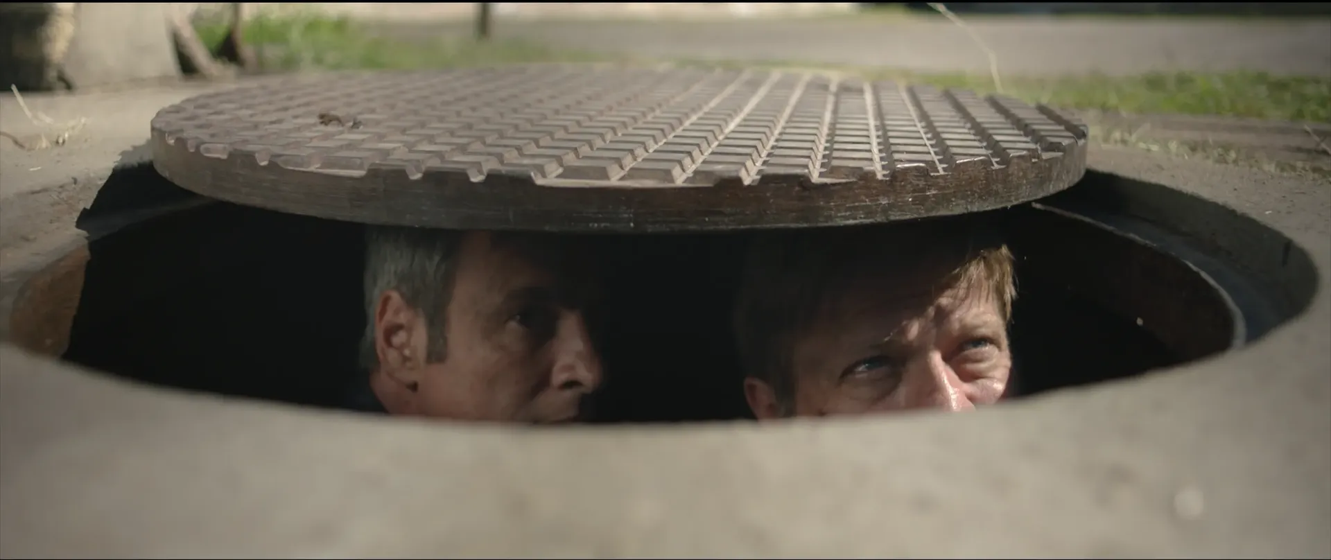 Two men peek out cautiously from beneath a partially lifted manhole cover, their faces lit by daylight as they look around from inside the sewer.