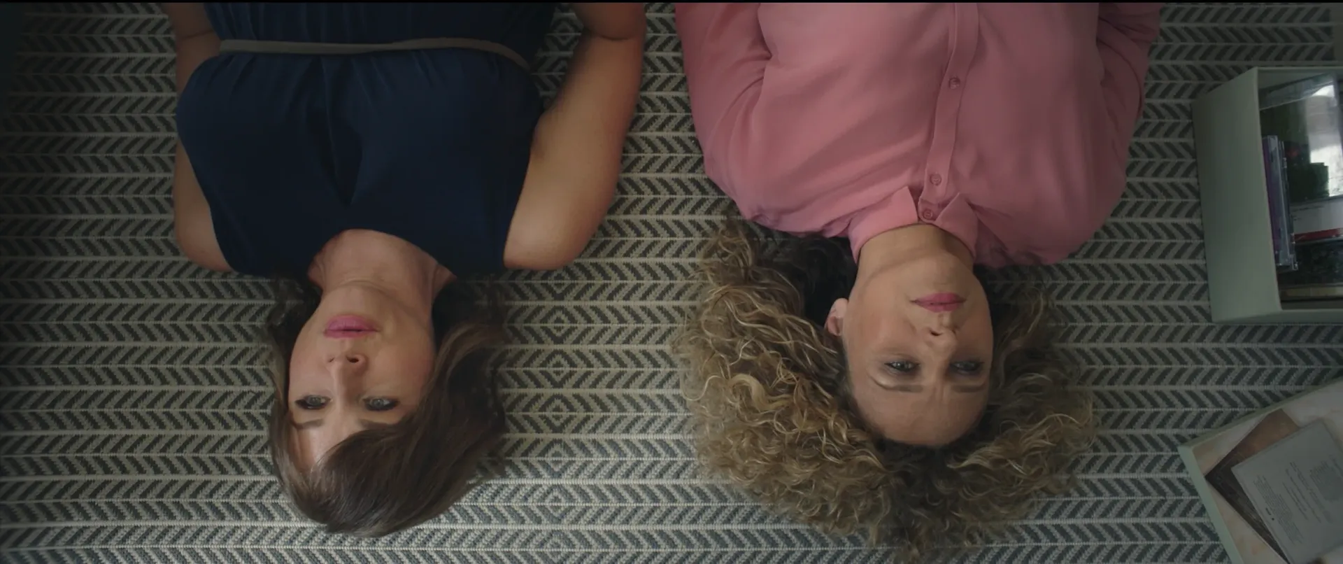 Two women lie side by side on a patterned carpet, viewed from above, one wearing a navy dress and the other a pink blouse, both staring upward with calm, thoughtful expressions.