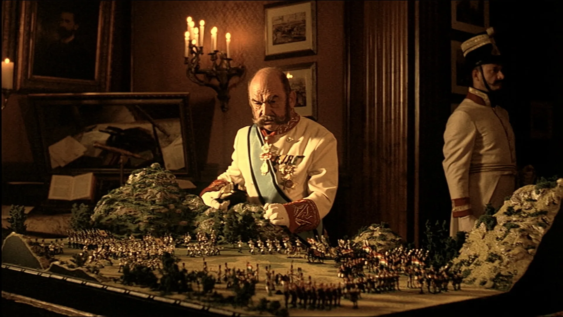 An older man in an ornate white military uniform studies a large miniature battlefield diorama in a dimly lit, antique-style room, with a uniformed guard standing in the background.
