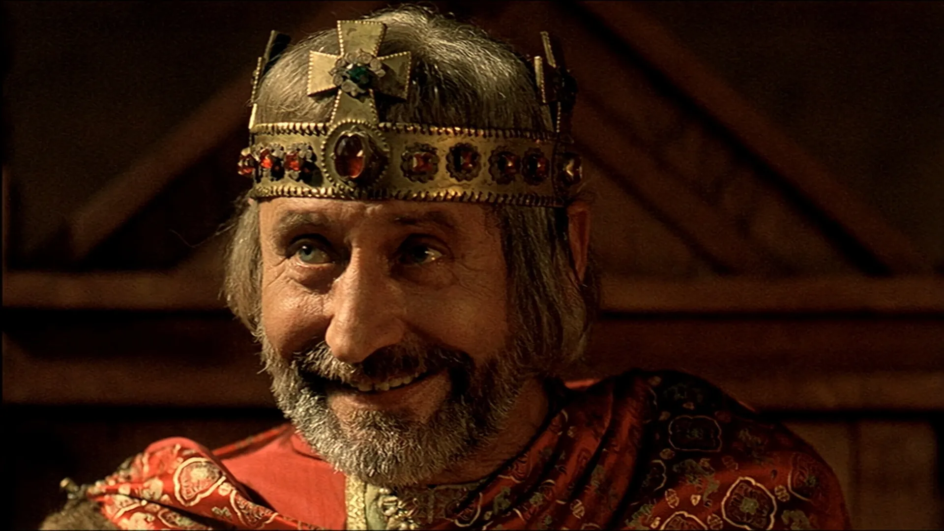 Smiling bearded man wearing an ornate golden crown decorated with red jewels, dressed in richly patterned medieval-style clothing inside a dimly lit hall.