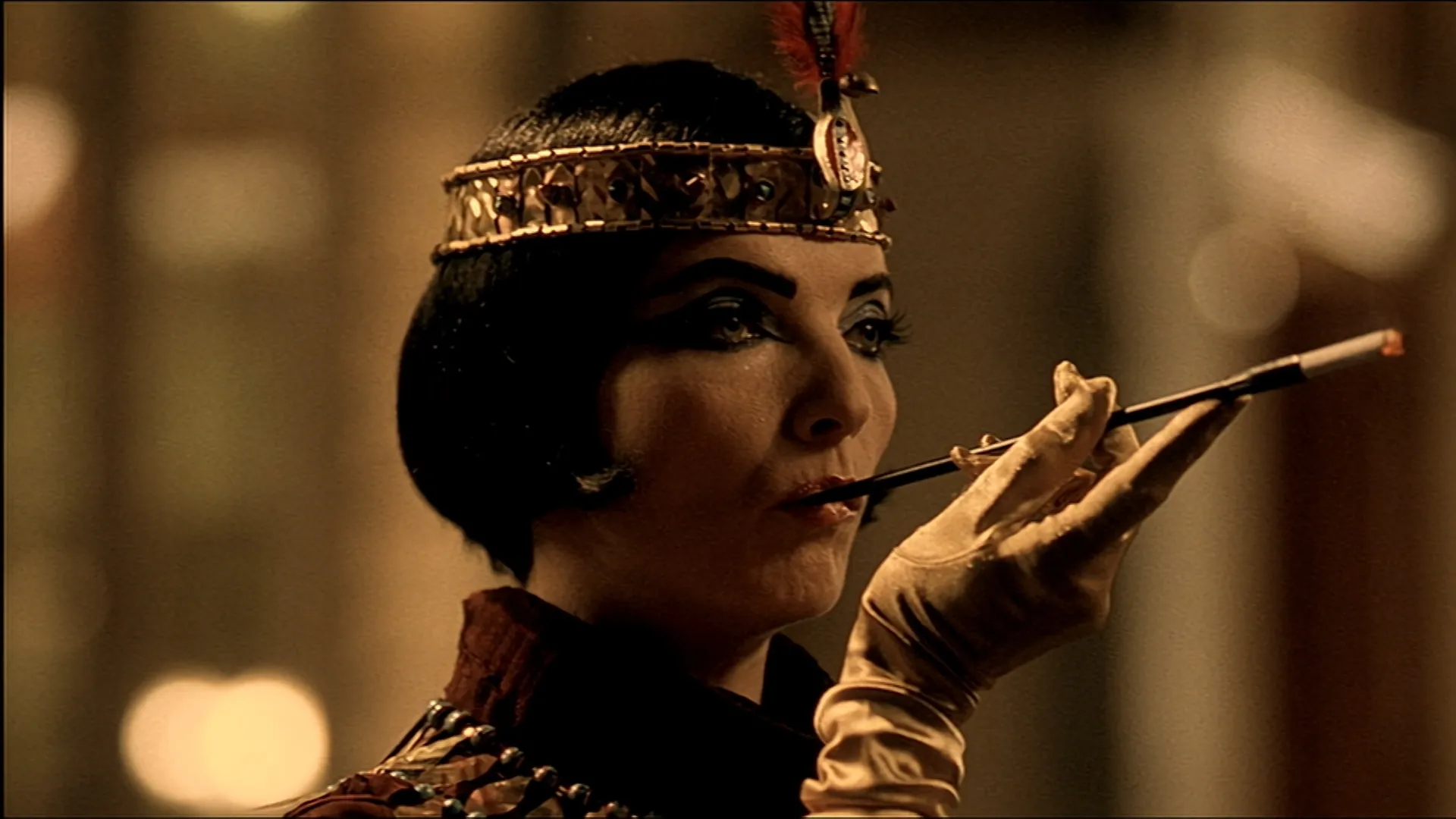 A woman dressed in ornate, vintage-style attire with dramatic black eye makeup and a jeweled headband smokes a cigarette from a long holder, her gloved hand lifted gracefully.