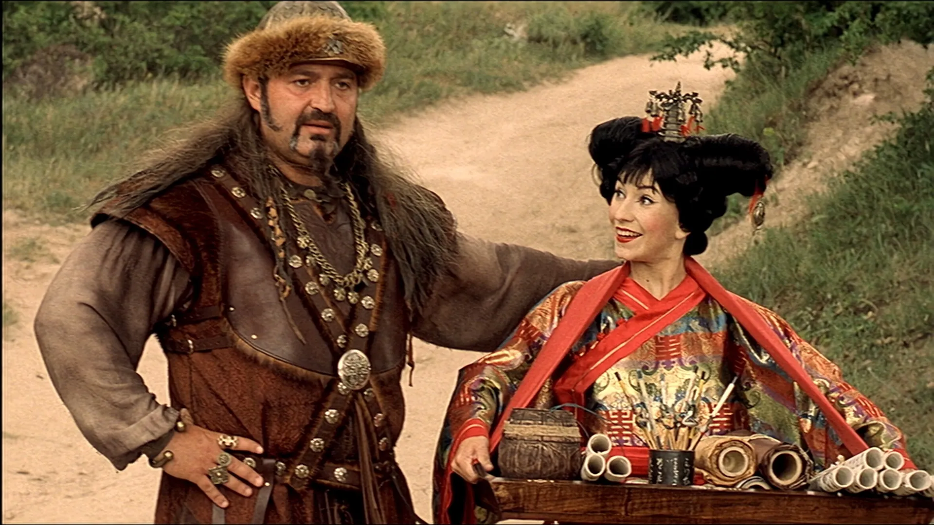 A bearded warrior in fur-trimmed leather armor stands beside a smiling woman dressed in an ornate, colorful robe with an elaborate hairstyle, both posed next to a wooden table filled with scrolls and writing tools on a dirt path surrounded by greenery.