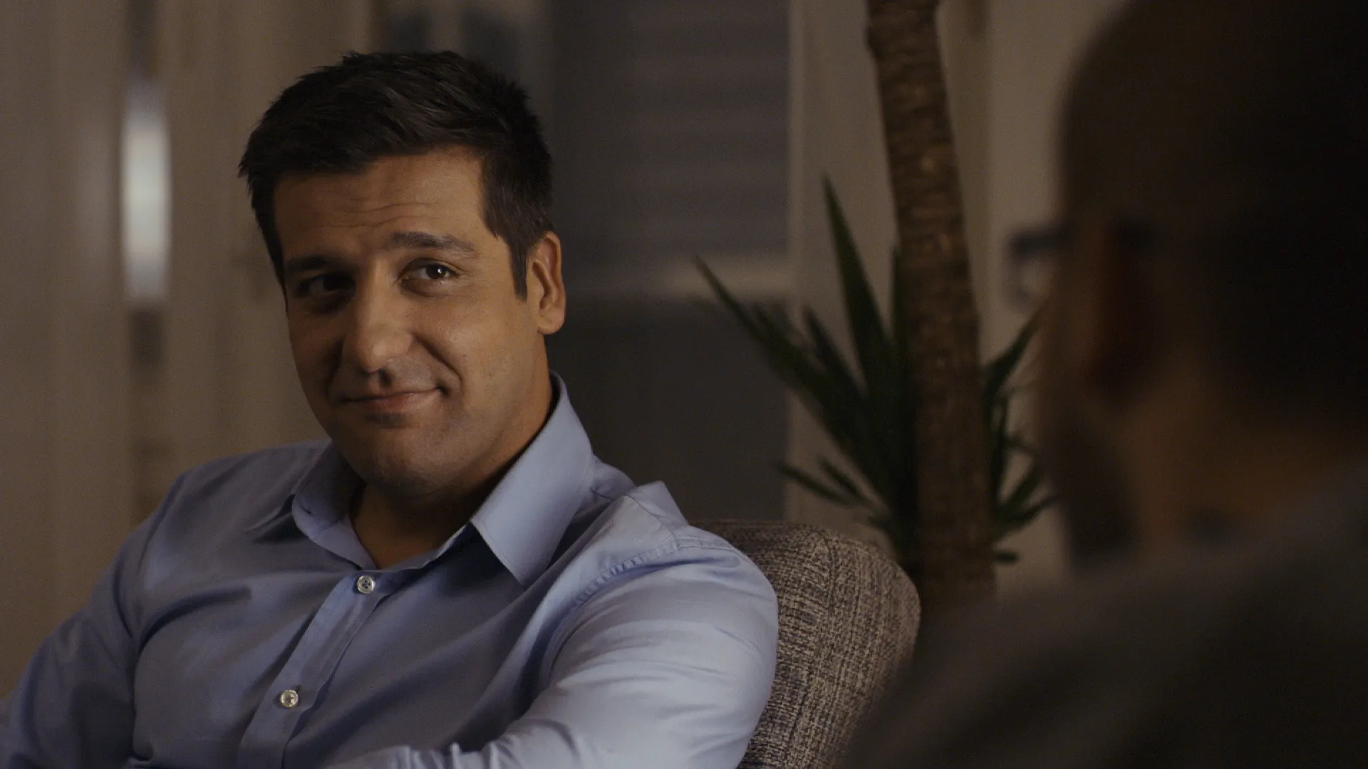 Man in a blue shirt sitting on a couch, giving a slight, knowing smile toward someone off-camera.