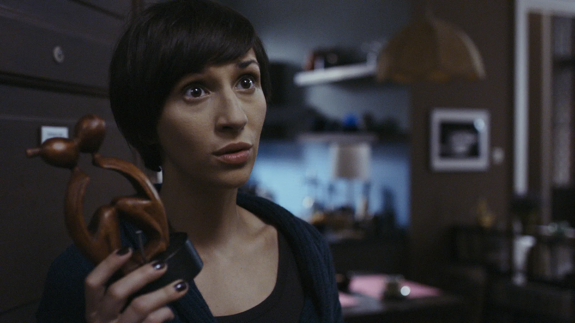 Woman with short dark hair holding a wooden figurine, looking upward with a concerned, questioning expression in a dimly lit room.