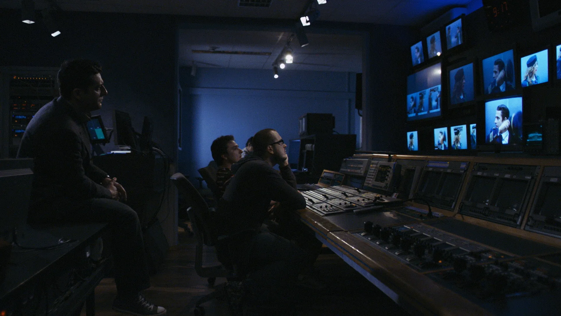 Three people sit in a dark control room, watching multiple blue-lit monitors on the wall while operating a large sound and video mixing console.