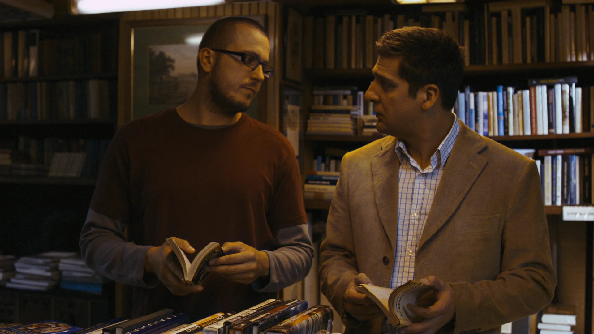 Two men stand in a cozy bookstore, flipping through books and looking at each other in mid-conversation.