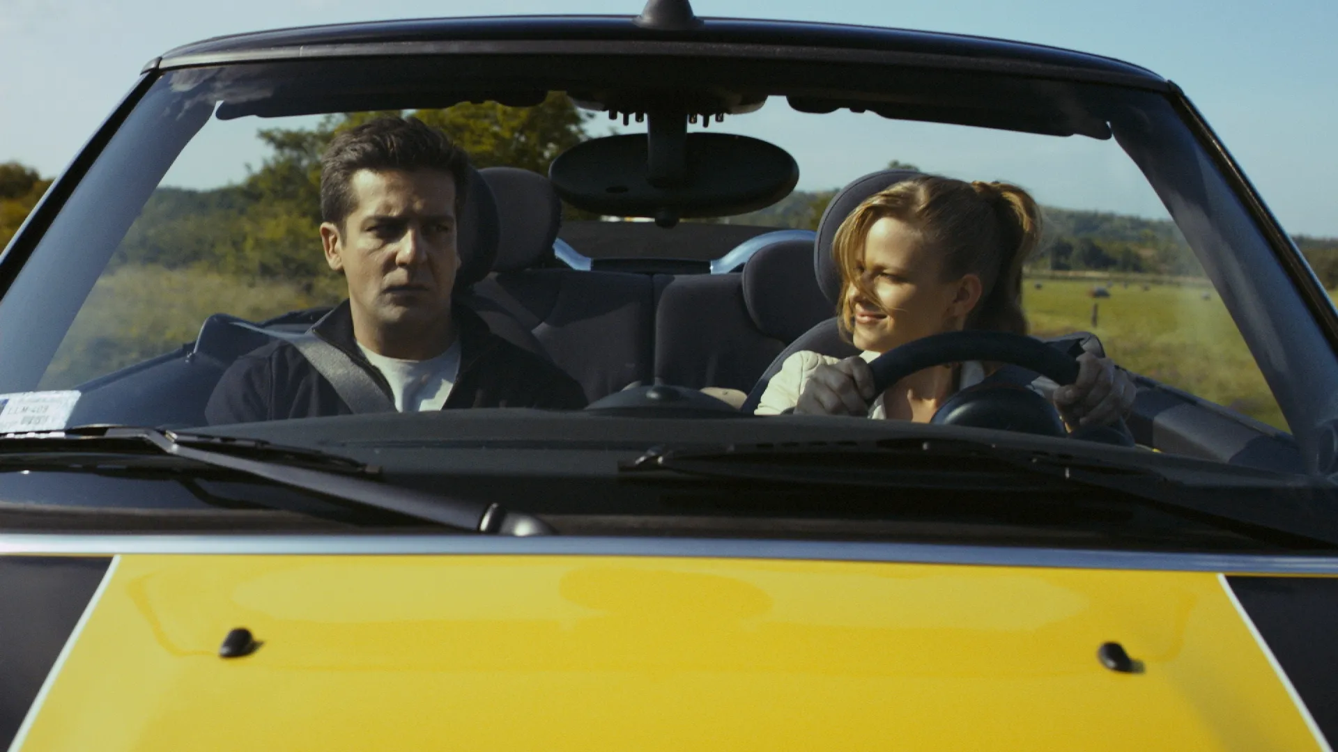Man and woman riding in a yellow convertible, with the woman smiling at the wheel while the man looks serious.