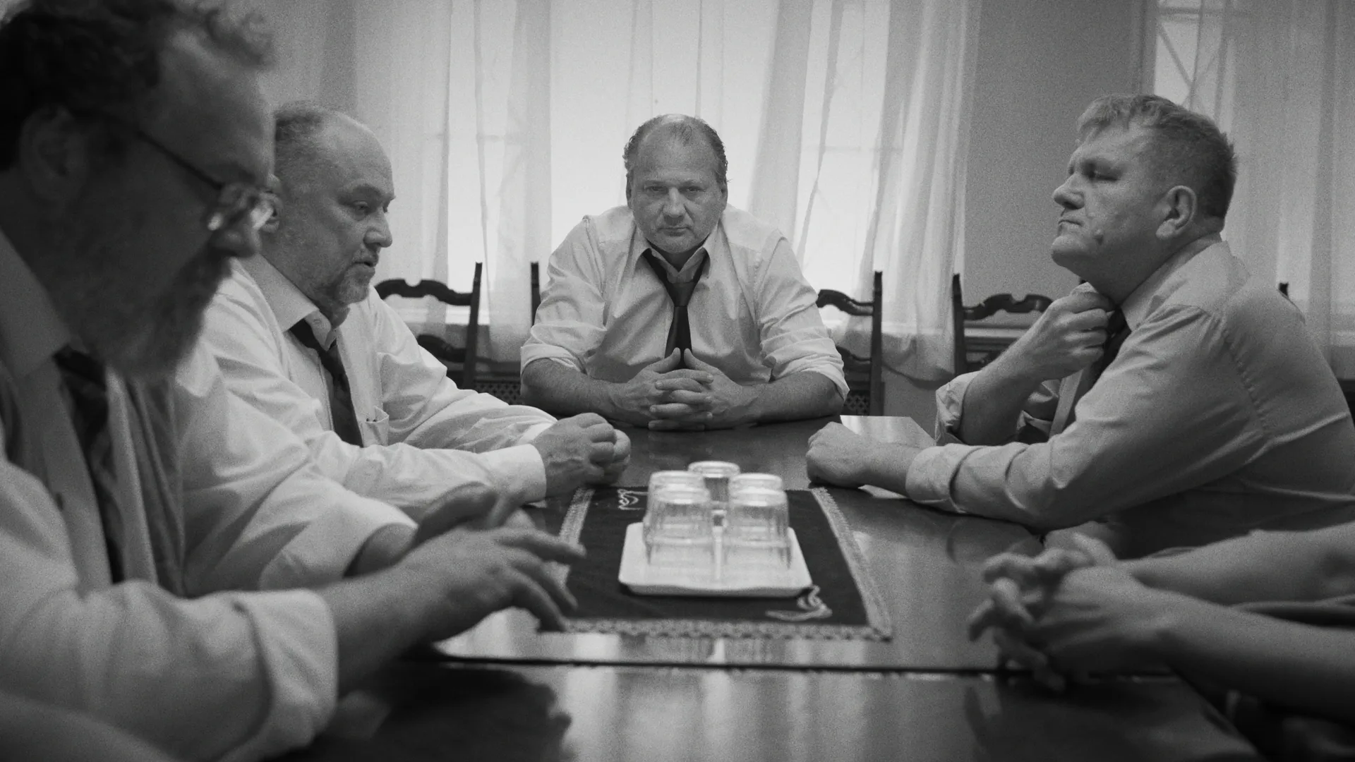 A group of men in shirts and loosened ties sit around a long table in a dimly lit room, facing each other in a tense meeting.