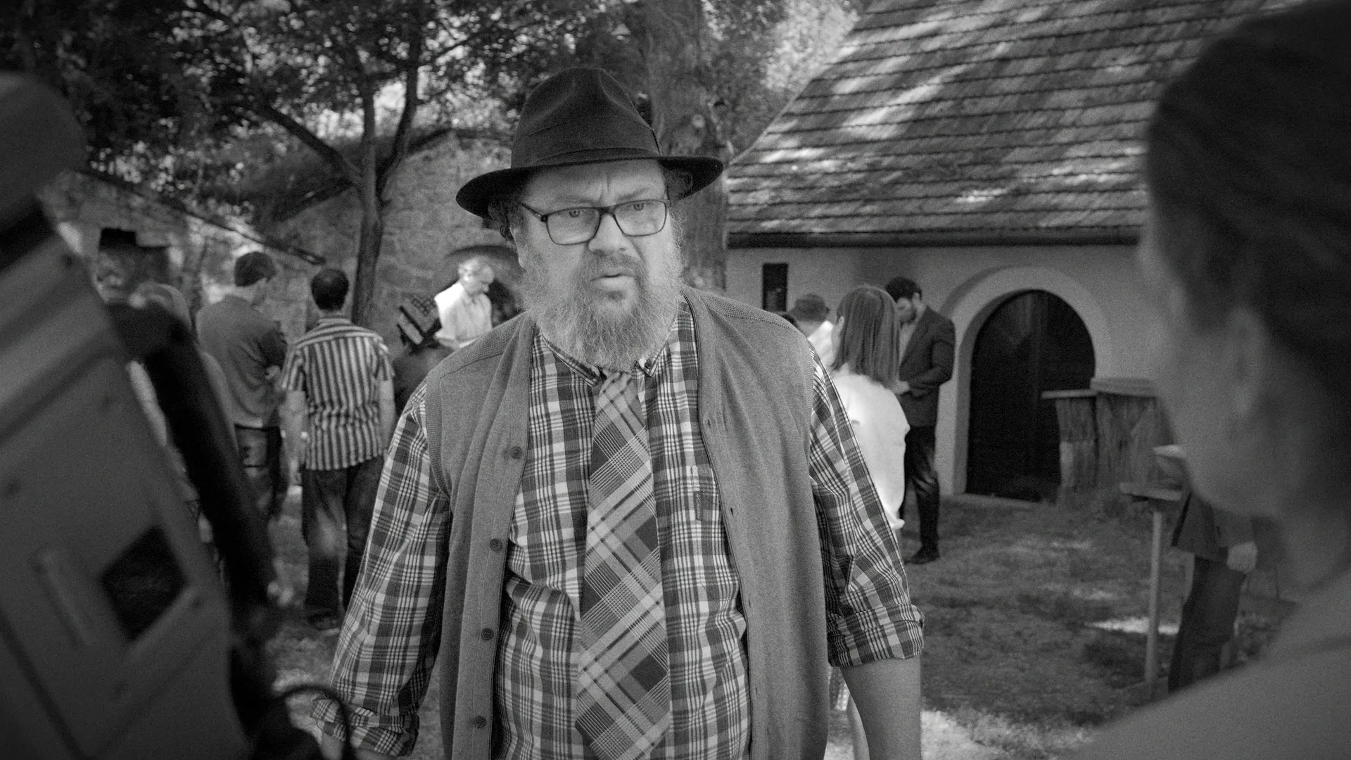 A bearded man in a hat and plaid shirt stands outdoors among a small gathering in a black-and-white village scene.