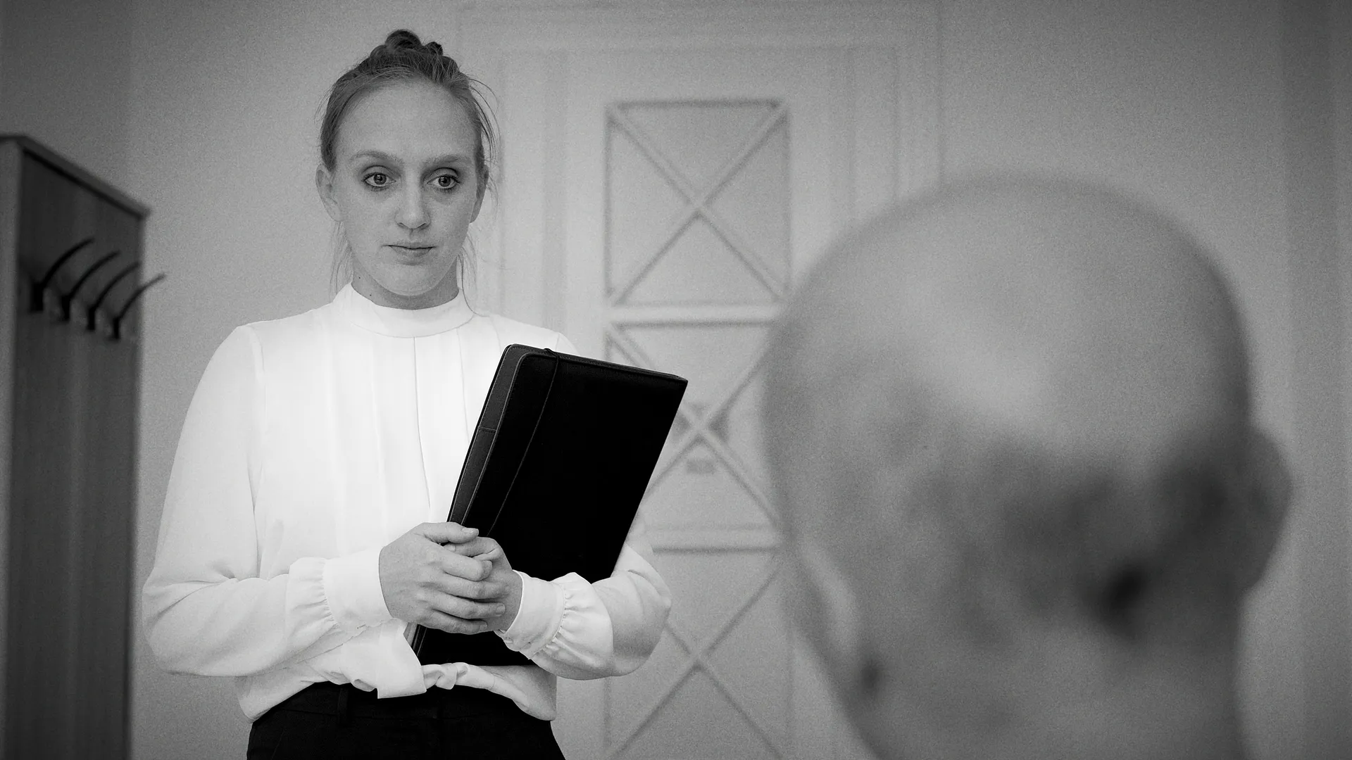A woman in a white blouse holds a folder while facing a seated bald man in a muted, black-and-white room.