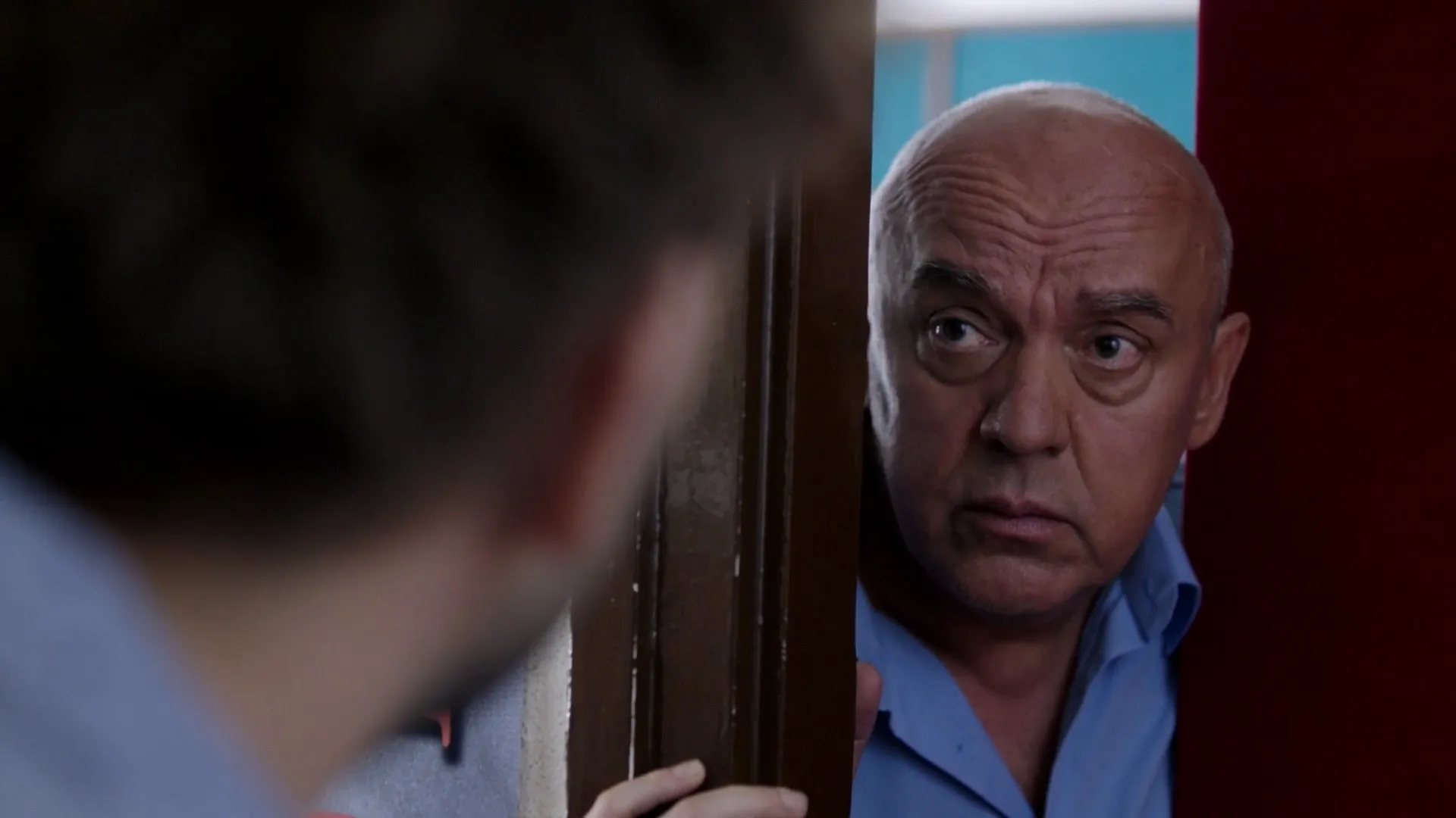 A middle-aged man with a serious, slightly concerned expression peers through a partially opened door, his hand resting on the frame. His blue shirt is visible, and he looks toward the blurred figure in the foreground, as if reacting to unexpected news or 