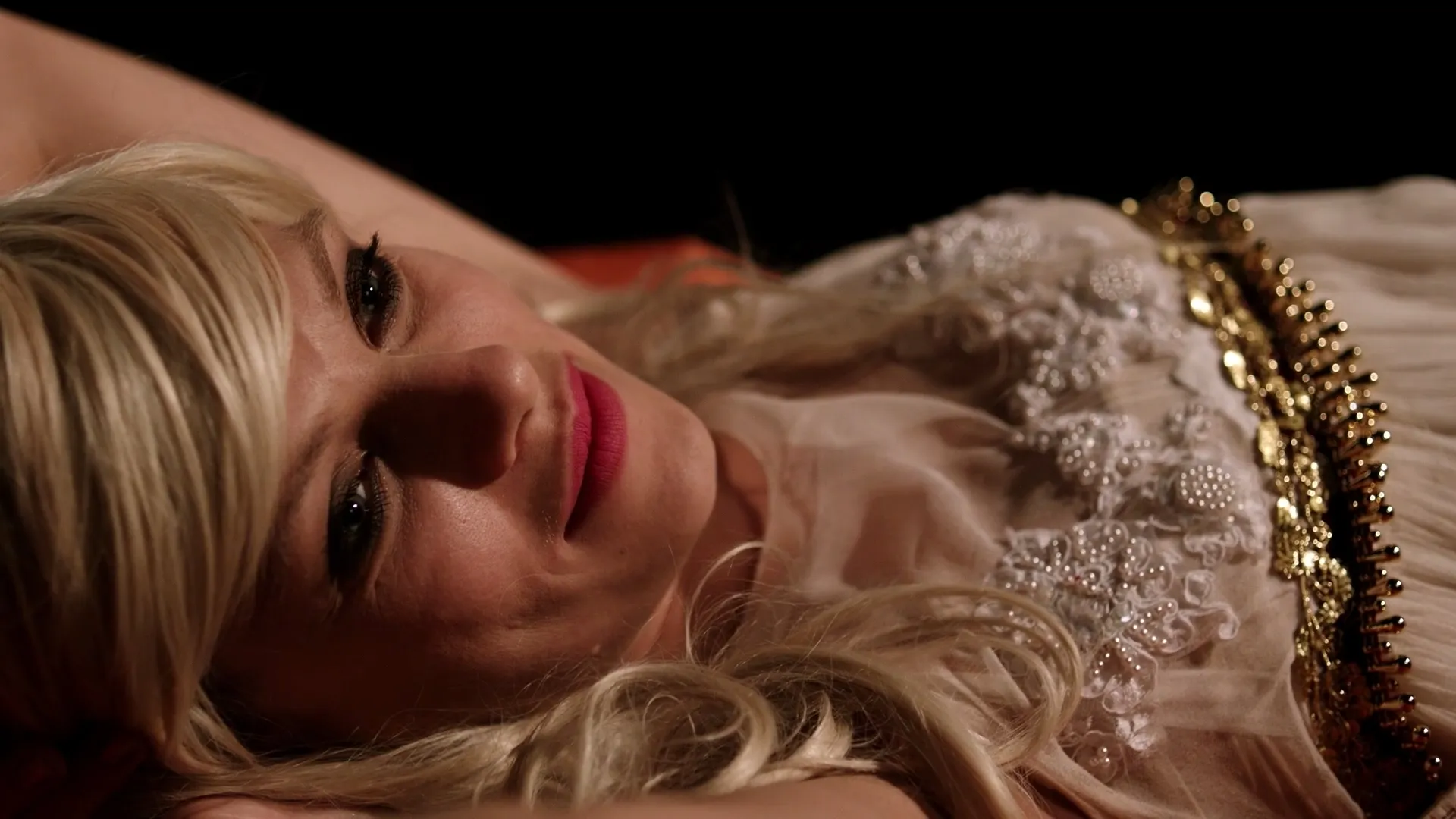 A blonde woman reclines in soft, warm light, wearing an ornate, beaded dress and gazing upward with a faint, calm smile.