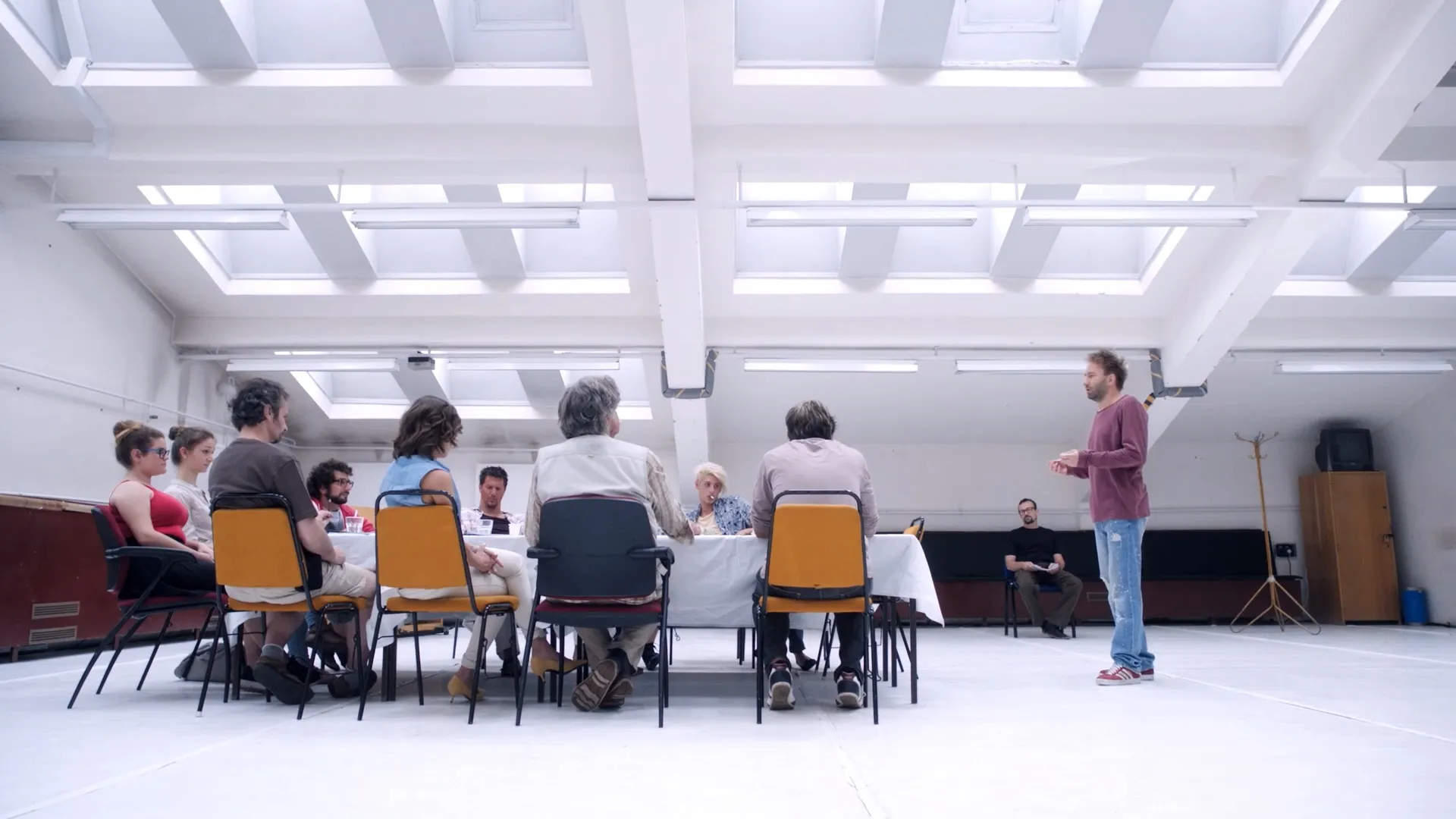 A bright rehearsal room where one man stands presenting to a long table of seated people, arranged like a committee or casting panel.